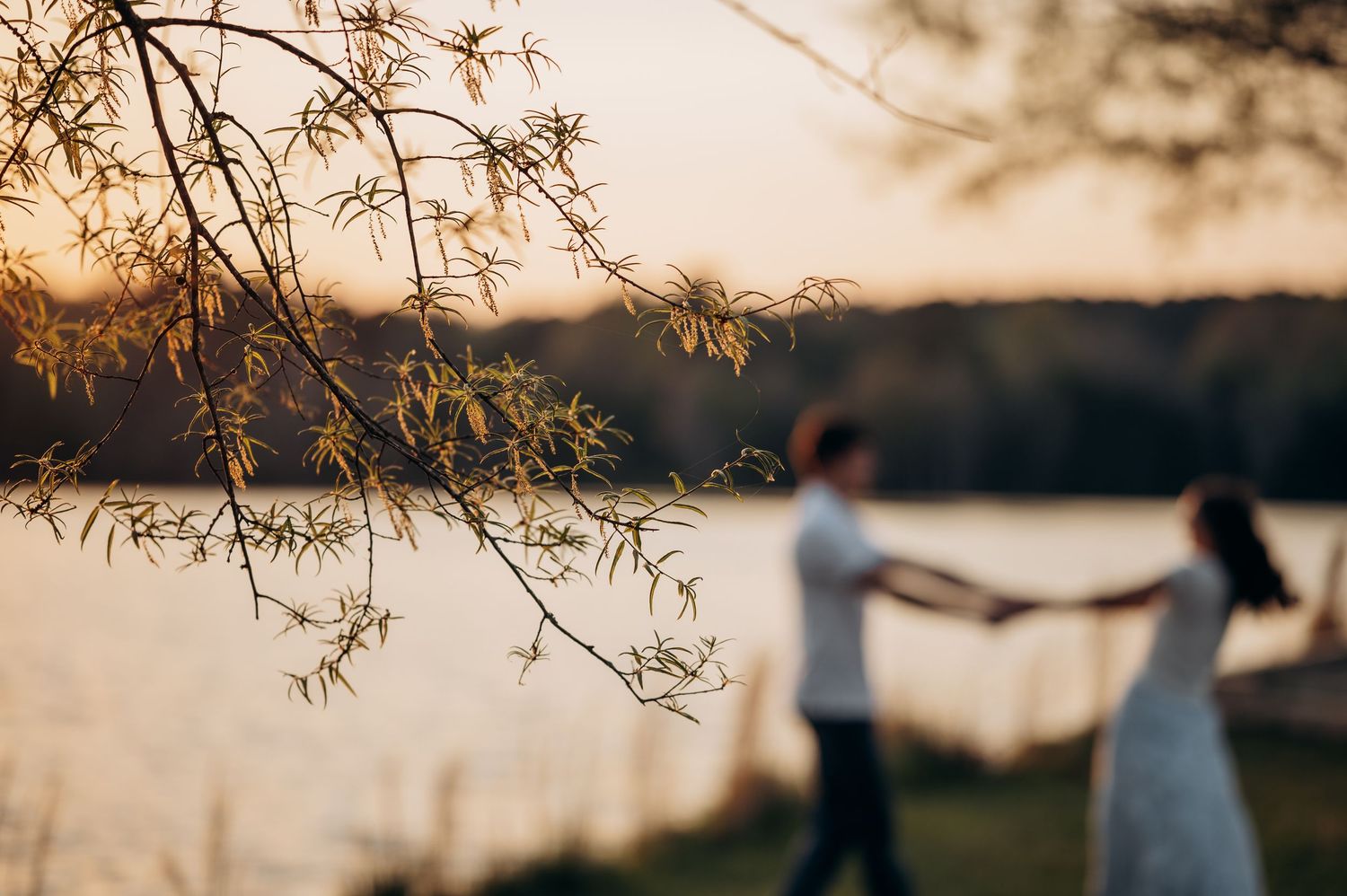 Couple holding hands by a serene lake at golden sunset with blurred tree branches in the foreground.
