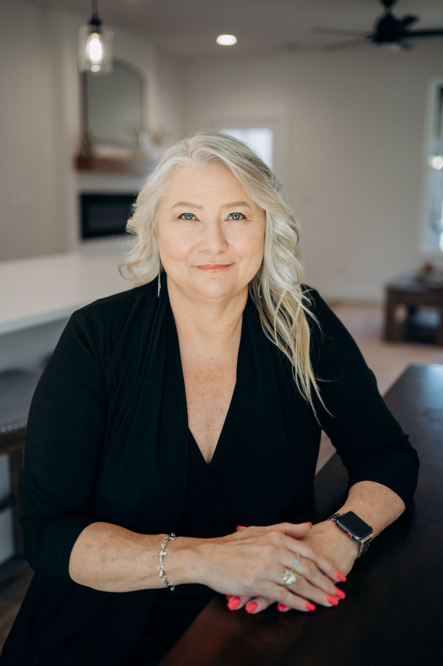 Smiling older woman with white hair in black top, seated at dark table in modern home interior.