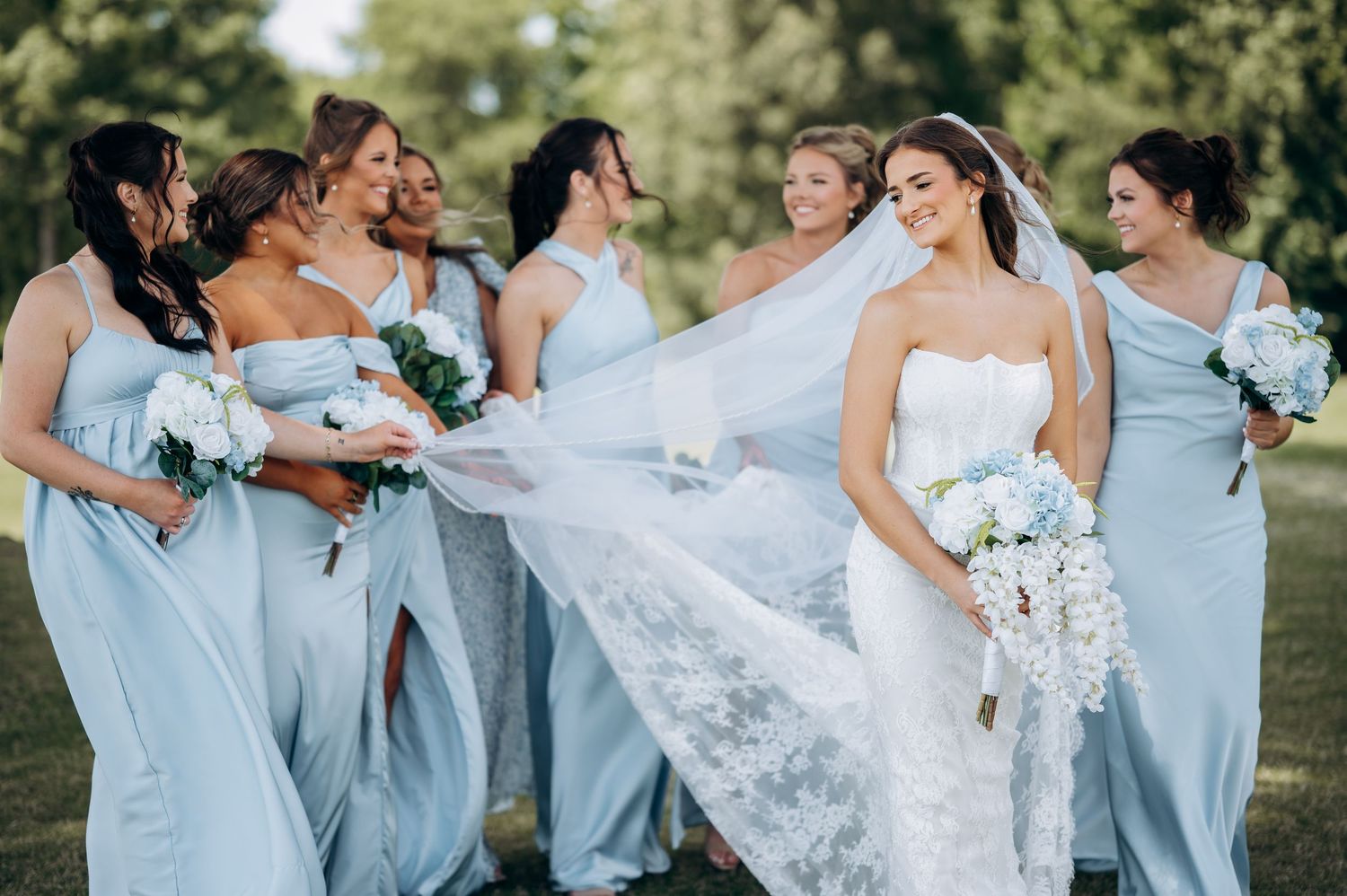Bride in white strapless gown with flowing veil surrounded by bridesmaids in light blue dresses holding white bouquets outdoors.