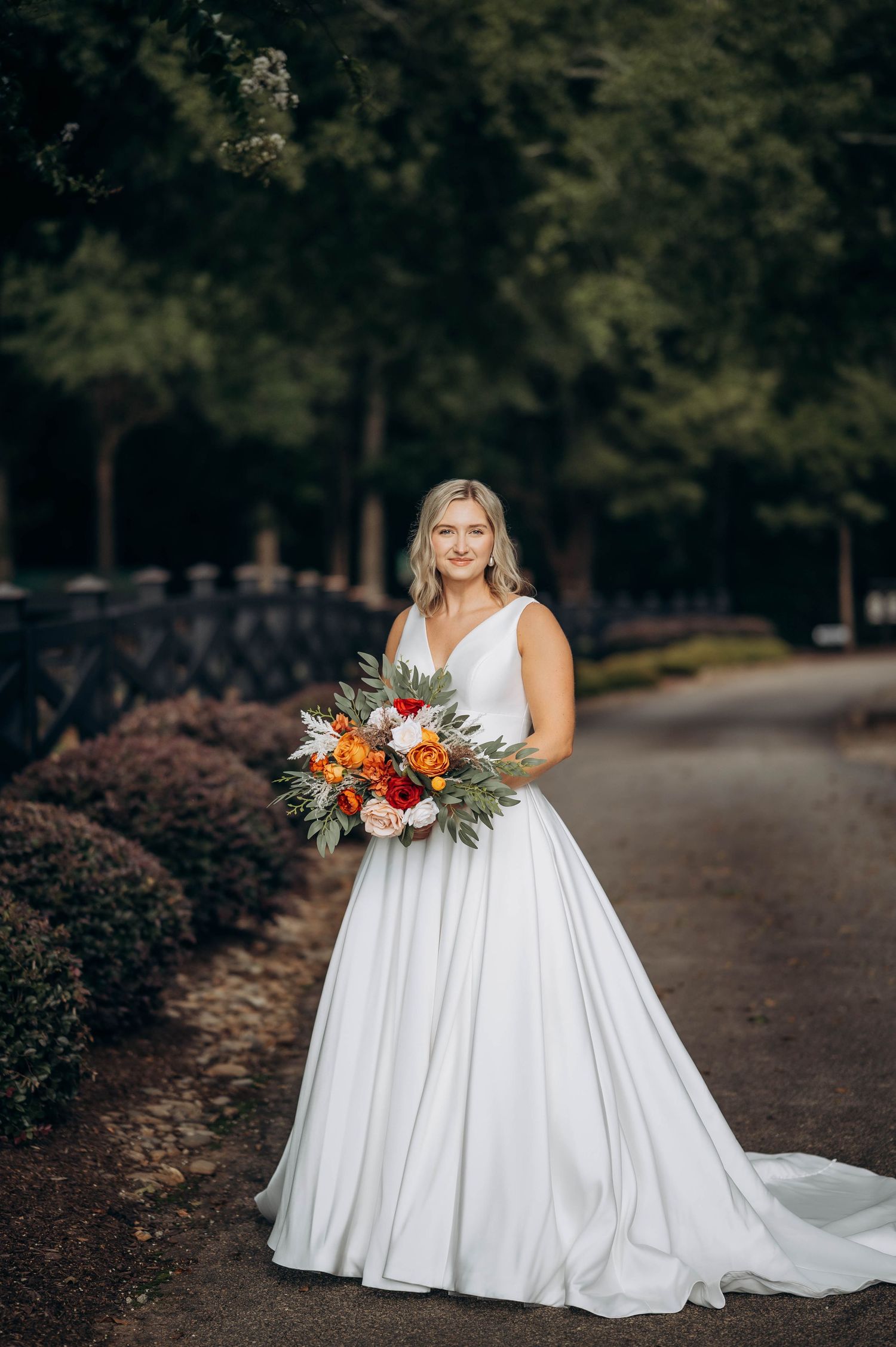 A bride in a flowing white gown holds a vibrant orange and white bouquet on a garden path.