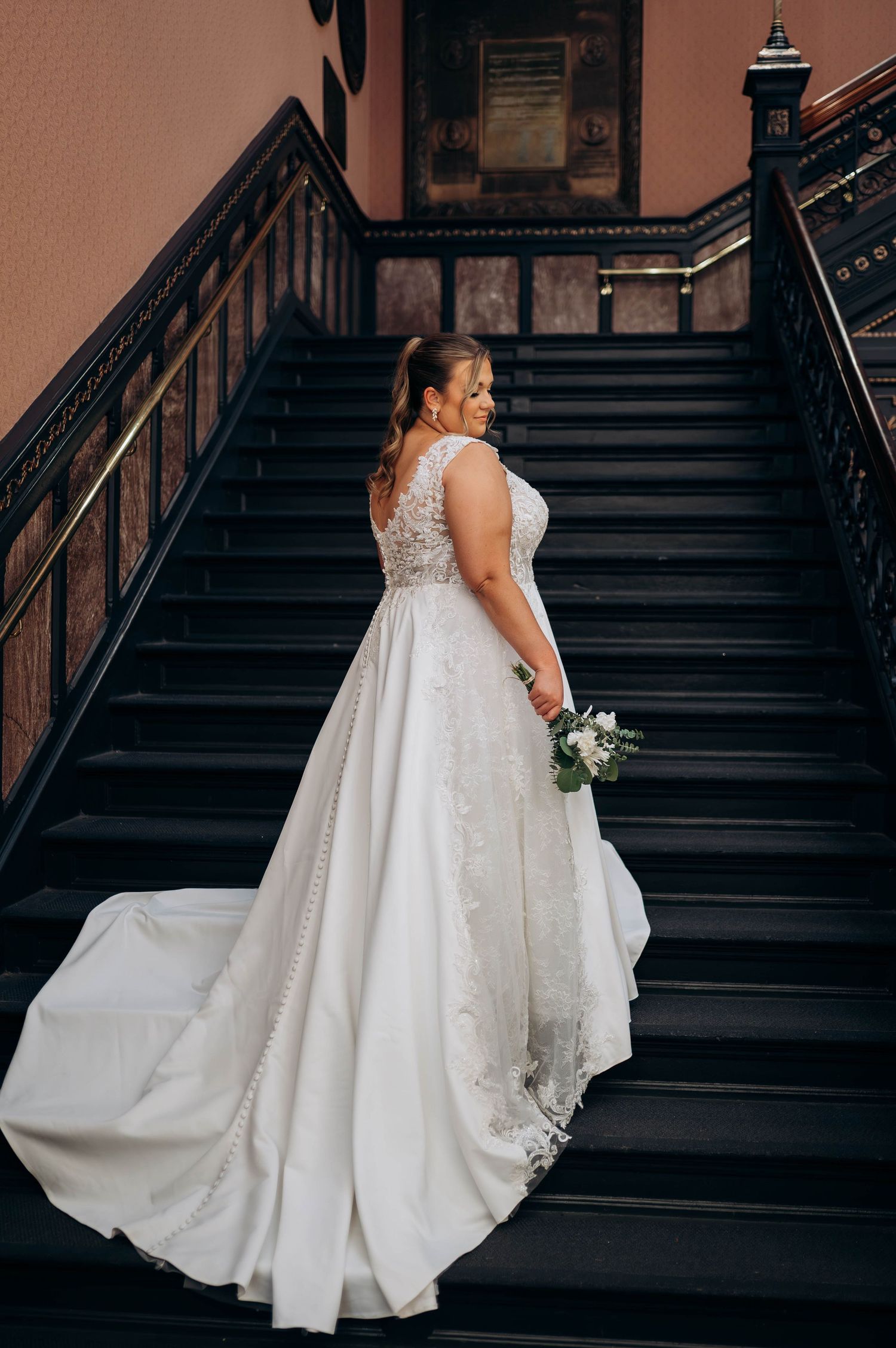 Flowing white wedding gown with train photographed on dark wooden staircase.
