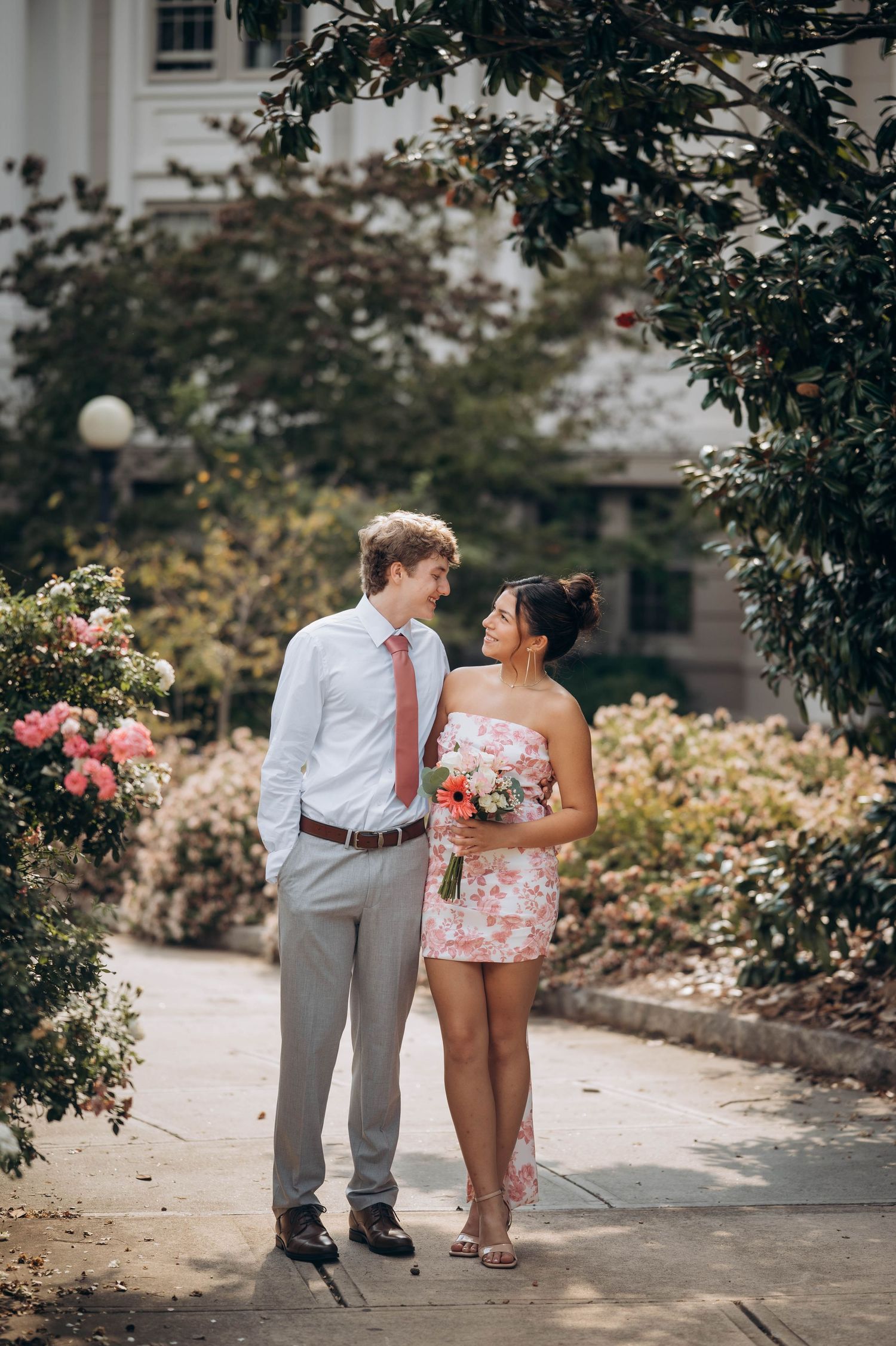 A sweet couple embracing on a garden walkway surrounded by pink roses and greenery.