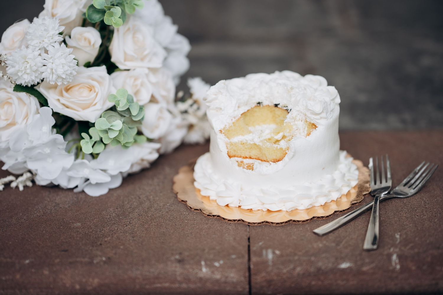 Sliced small white wedding cake beside a bouquet of white roses and two forks on a stone surface.