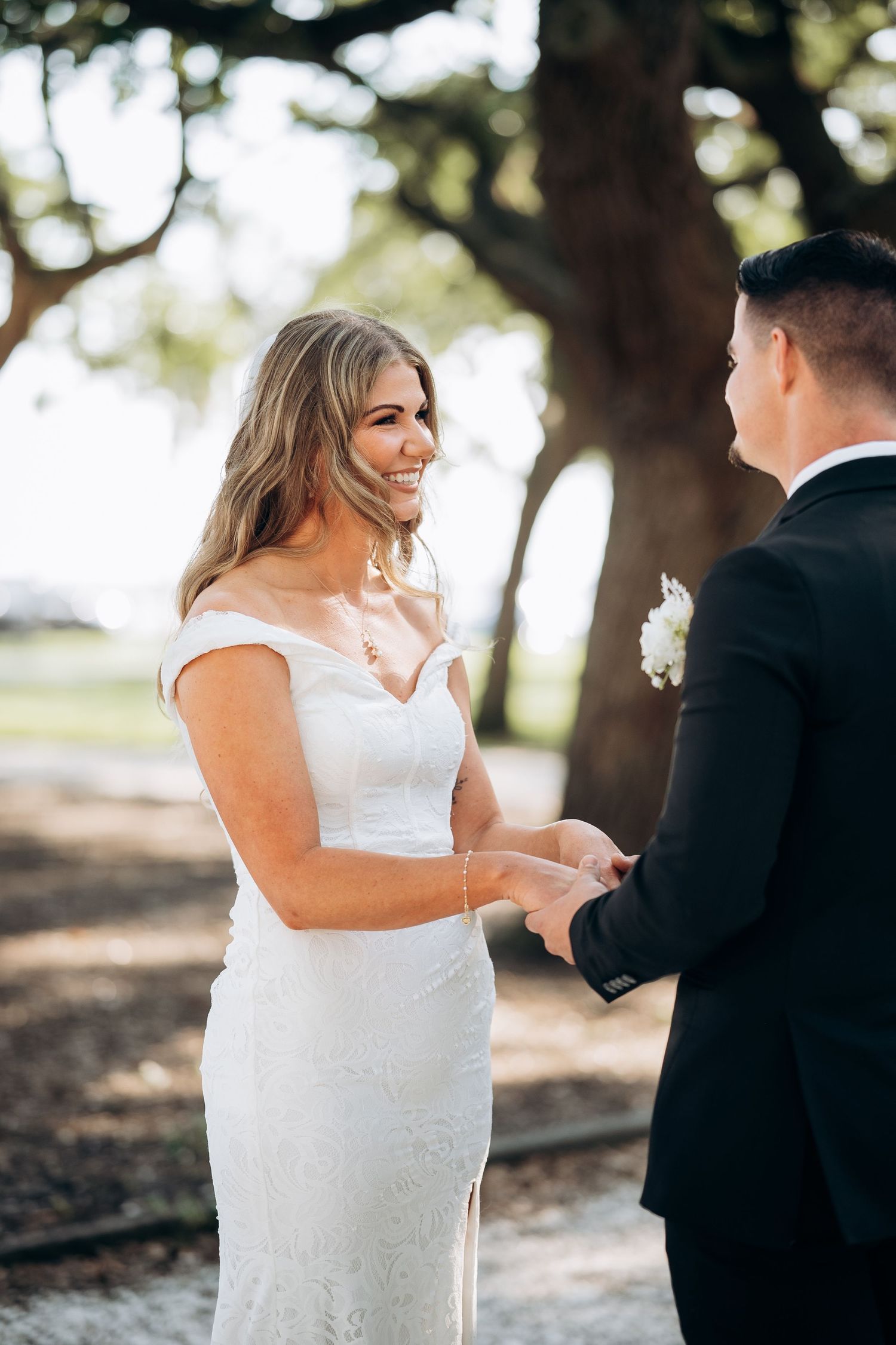 Smiling bride in white off-shoulder gown holds hands with groom in black suit during outdoor wedding ceremony.