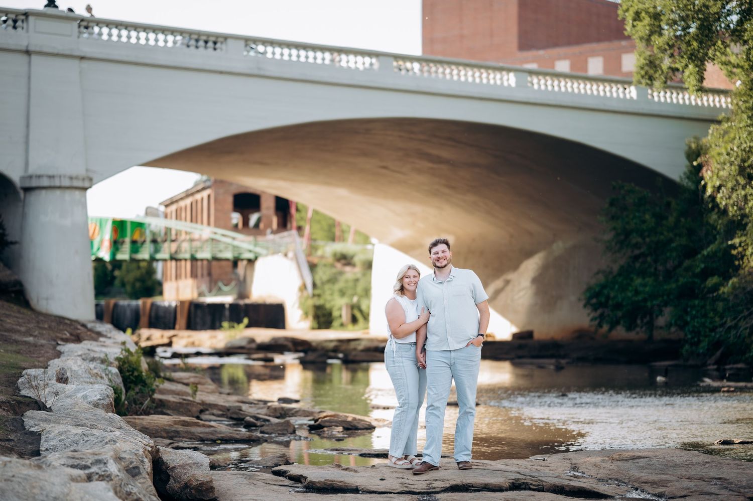 Couple standing by a river under a stone bridge in an urban setting, smiling together on a sunny day.