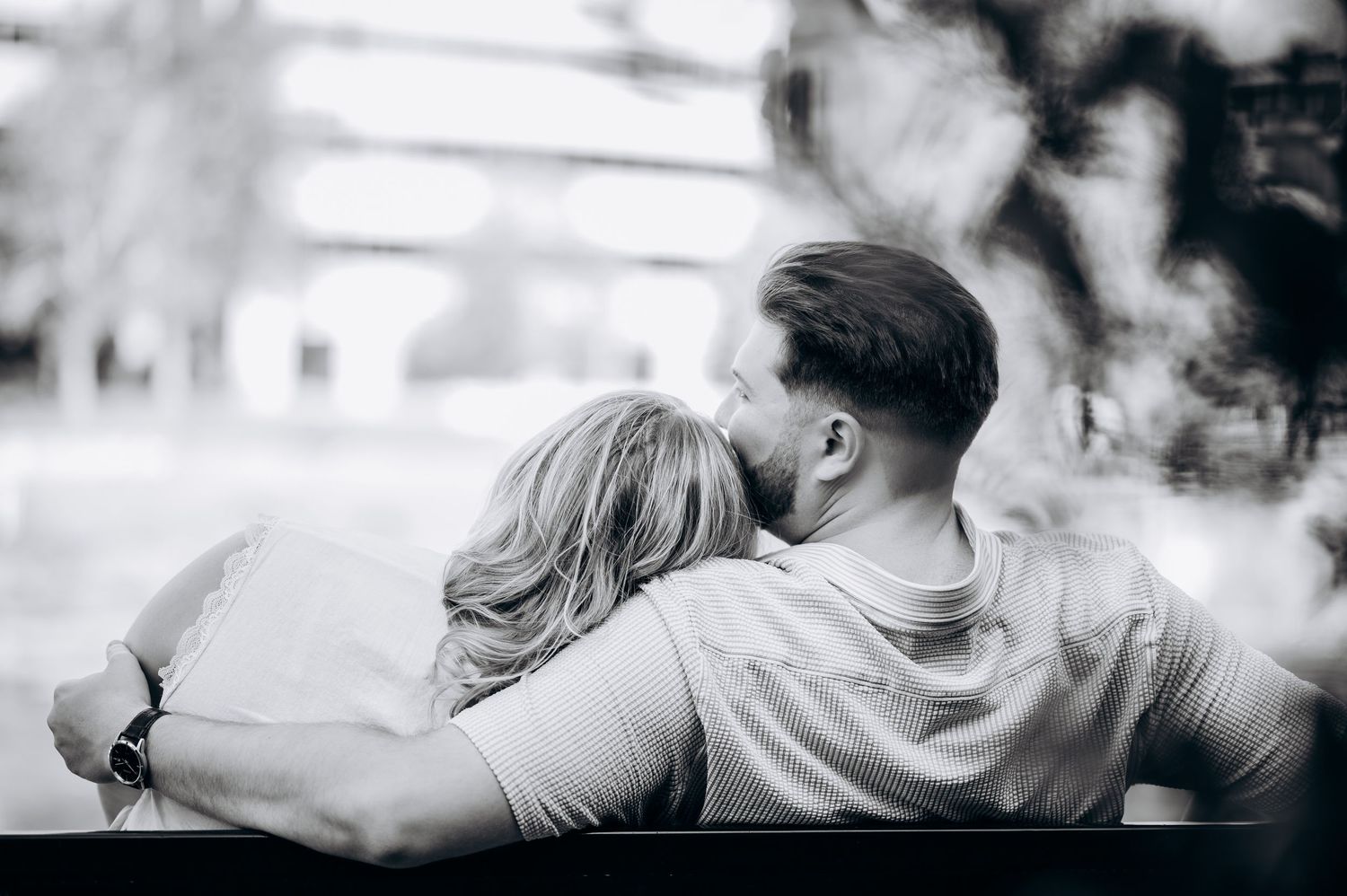 Black and white photo of a couple sitting close on a bench, man lovingly embracing woman outdoors.