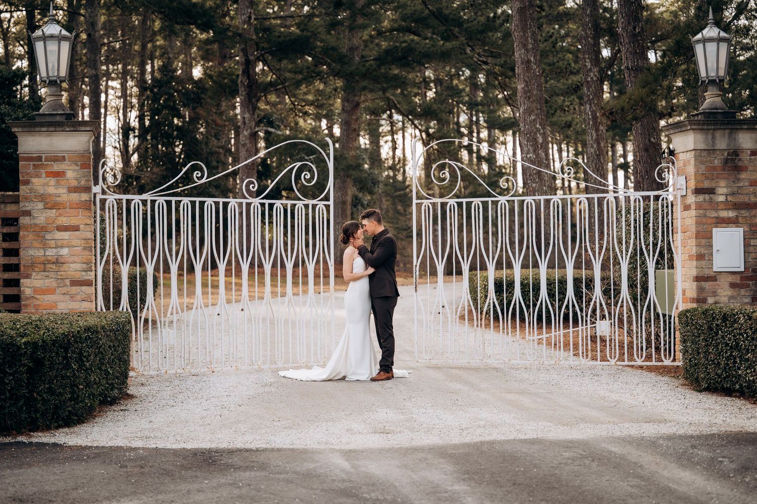 Bride and groom share a romantic kiss in front of elegant white iron gates surrounded by tall trees.