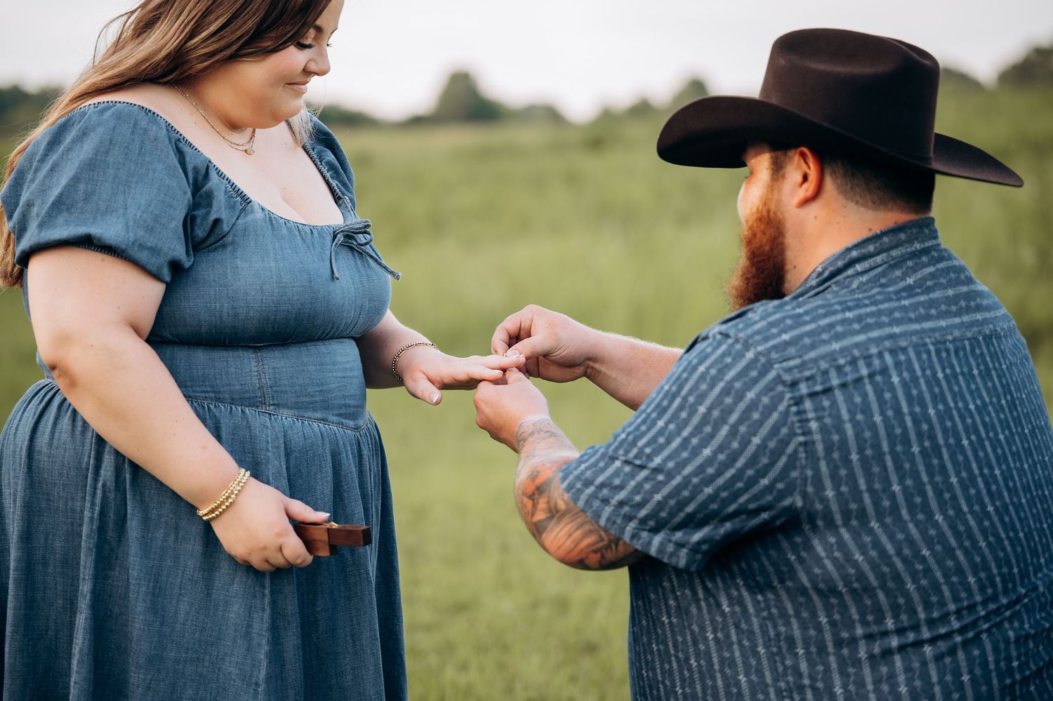 Man in cowboy hat kneeling to place a ring on a woman in a blue dress during an outdoor proposal.