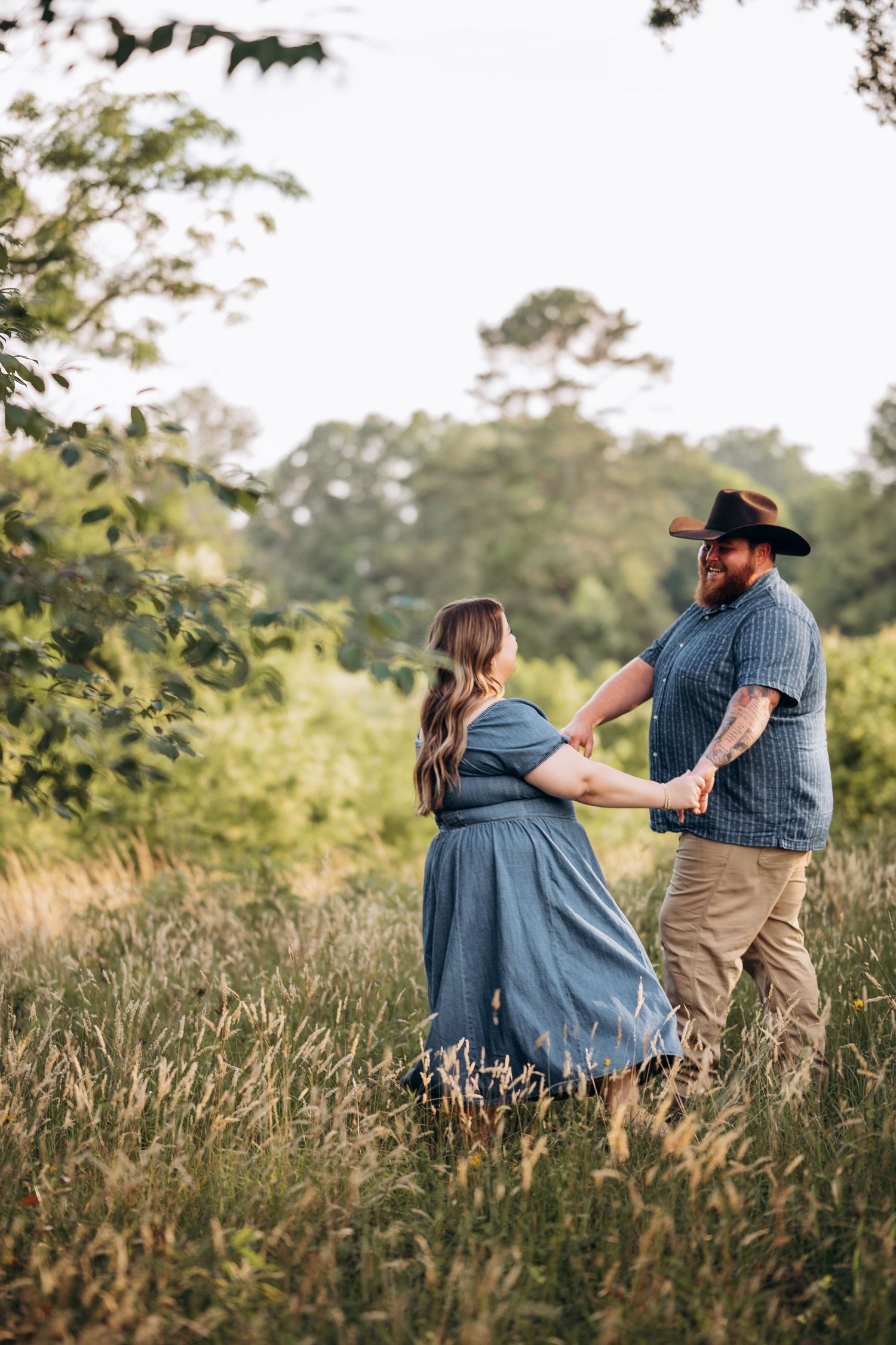 Couple dancing and holding hands in a tall grass meadow with green trees in the background during a sunny outdoor shoot.