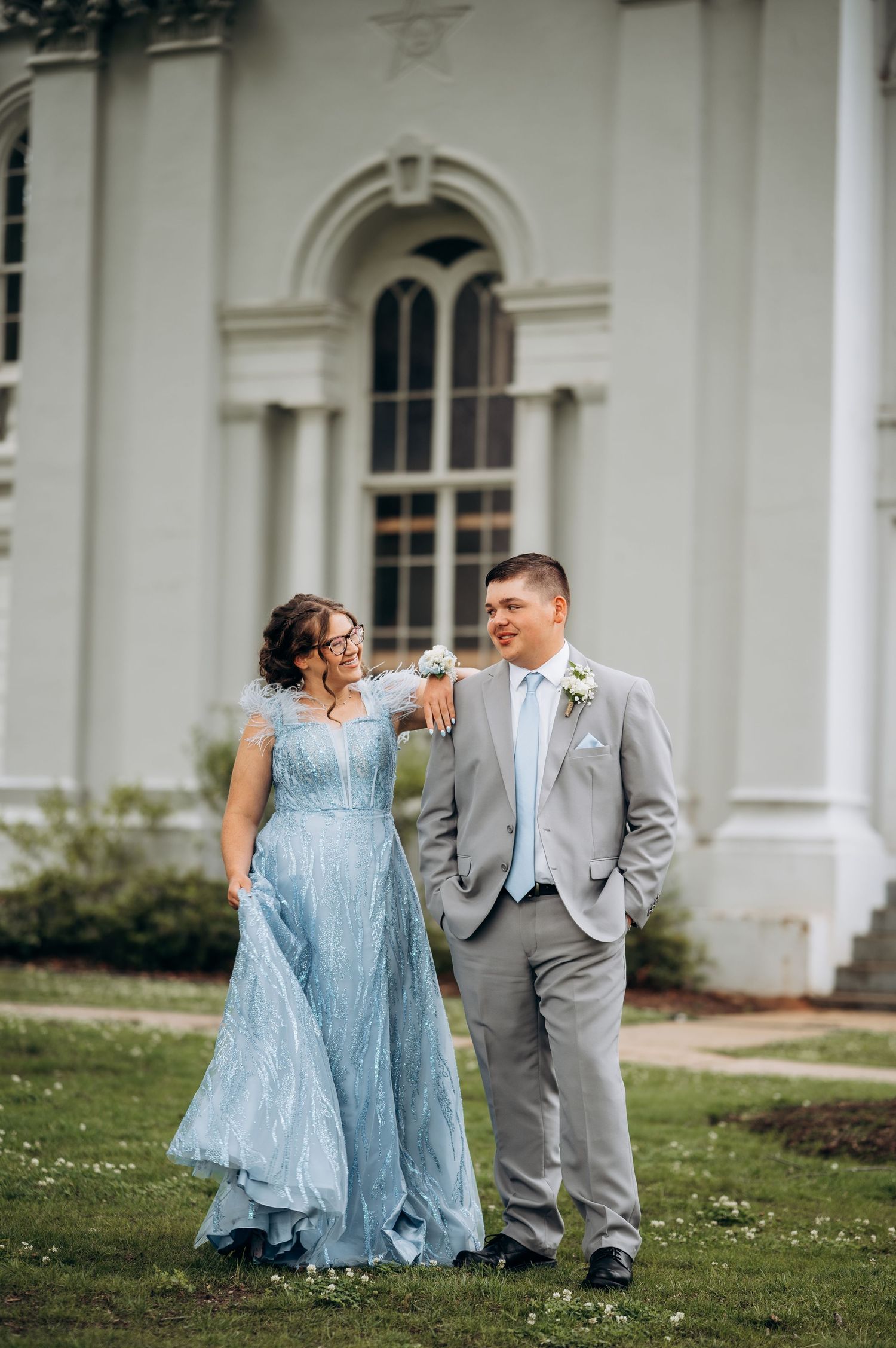 Couple in blue gown and gray suit walking together outside a white historic building for prom or wedding.