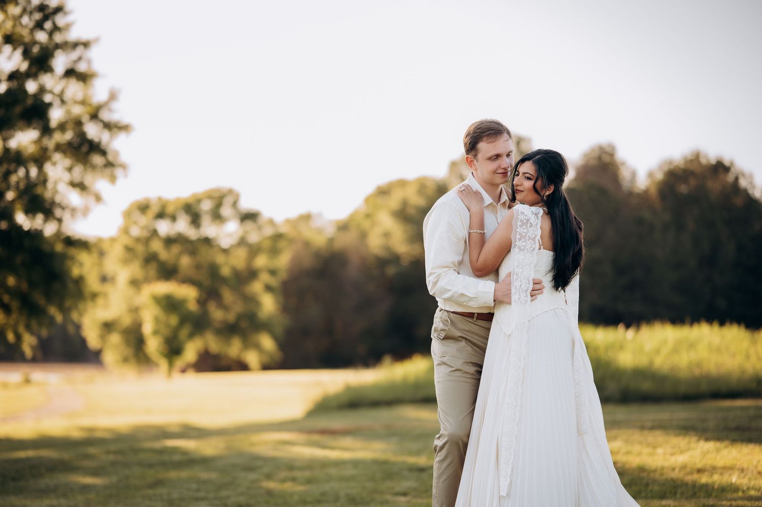 A couple shares a romantic embrace in a sunlit outdoor wedding setting surrounded by lush green trees and fields.