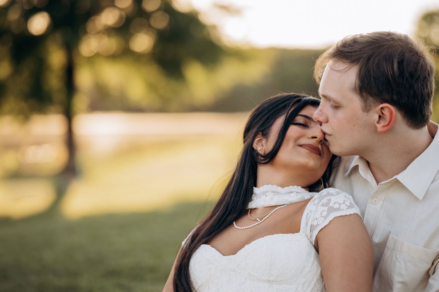 Groom kissing bride on cheek outdoors at sunset, bride in white lace dress smiling, romantic wedding portrait.