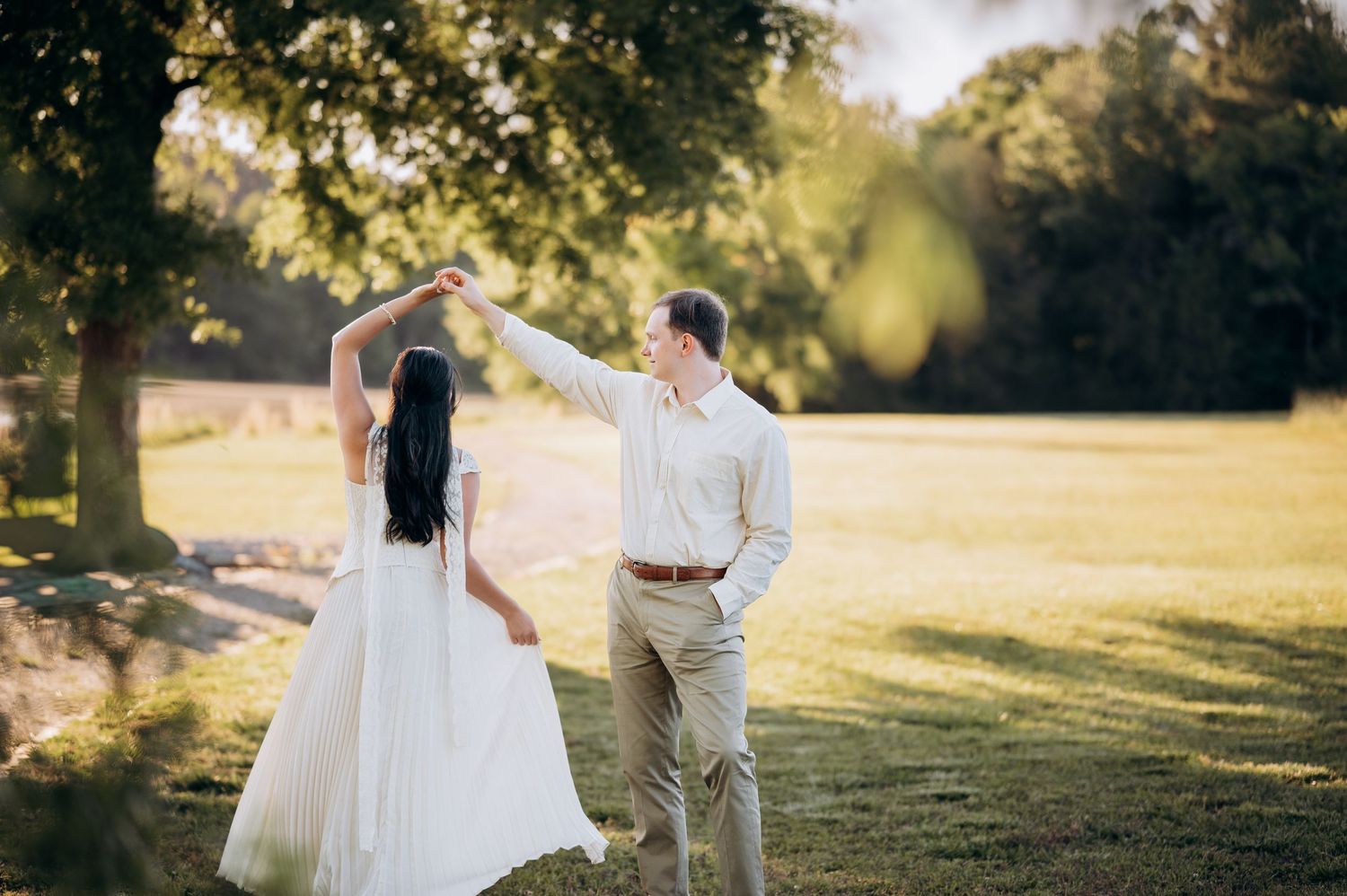 Couple dancing outdoors on green lawn, bride in white gown twirling under groom's hand in golden sunlight.