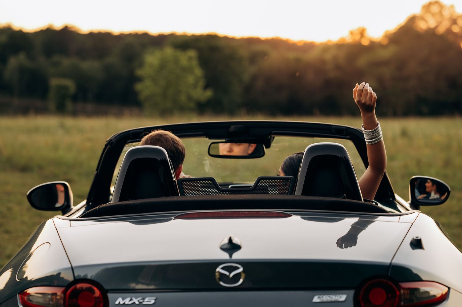 Person raising fist while riding in a silver Mazda MX-5 Miata convertible through a scenic countryside at sunset.