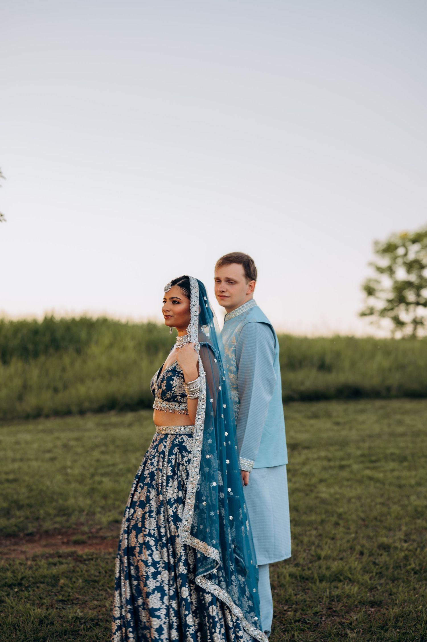 Couple in coordinating blue Indian wedding attire standing back-to-back in a green field at dusk.