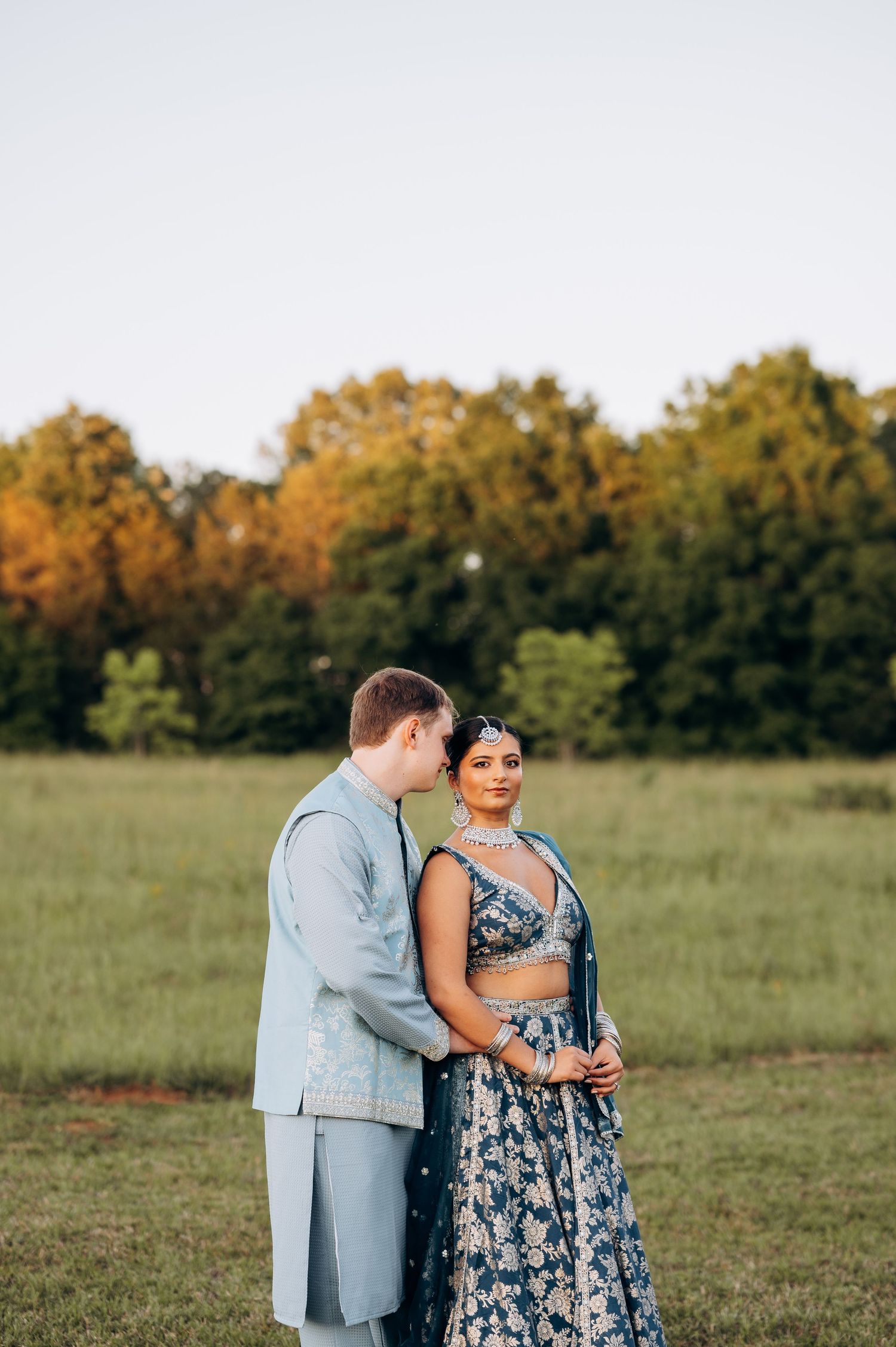 Couple in matching blue outfits pose in green field at sunset, woman in Indian lehenga, man in light blue suit.