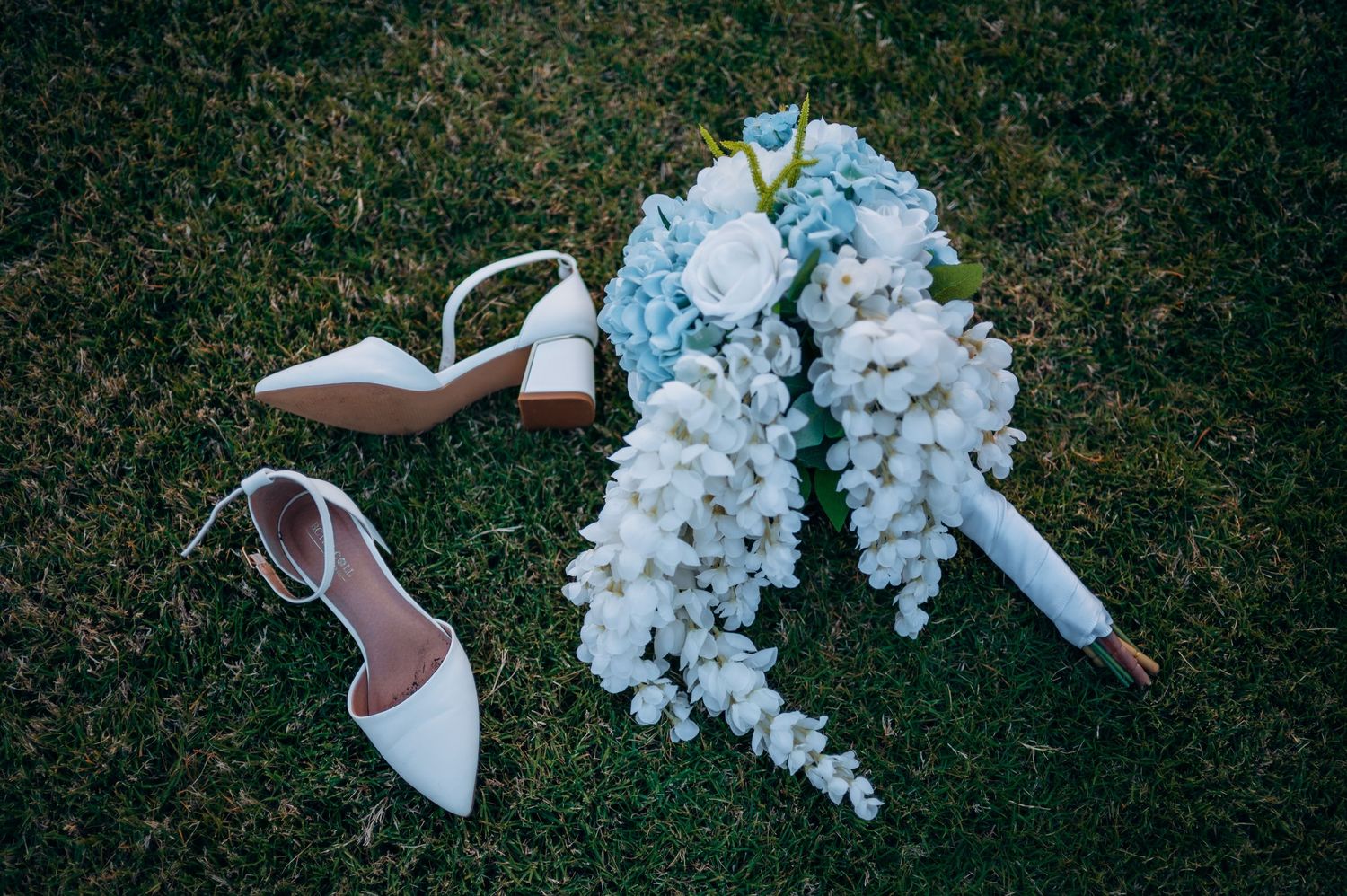 White bridal heels and cascading blue hydrangea bouquet arranged on green grass.