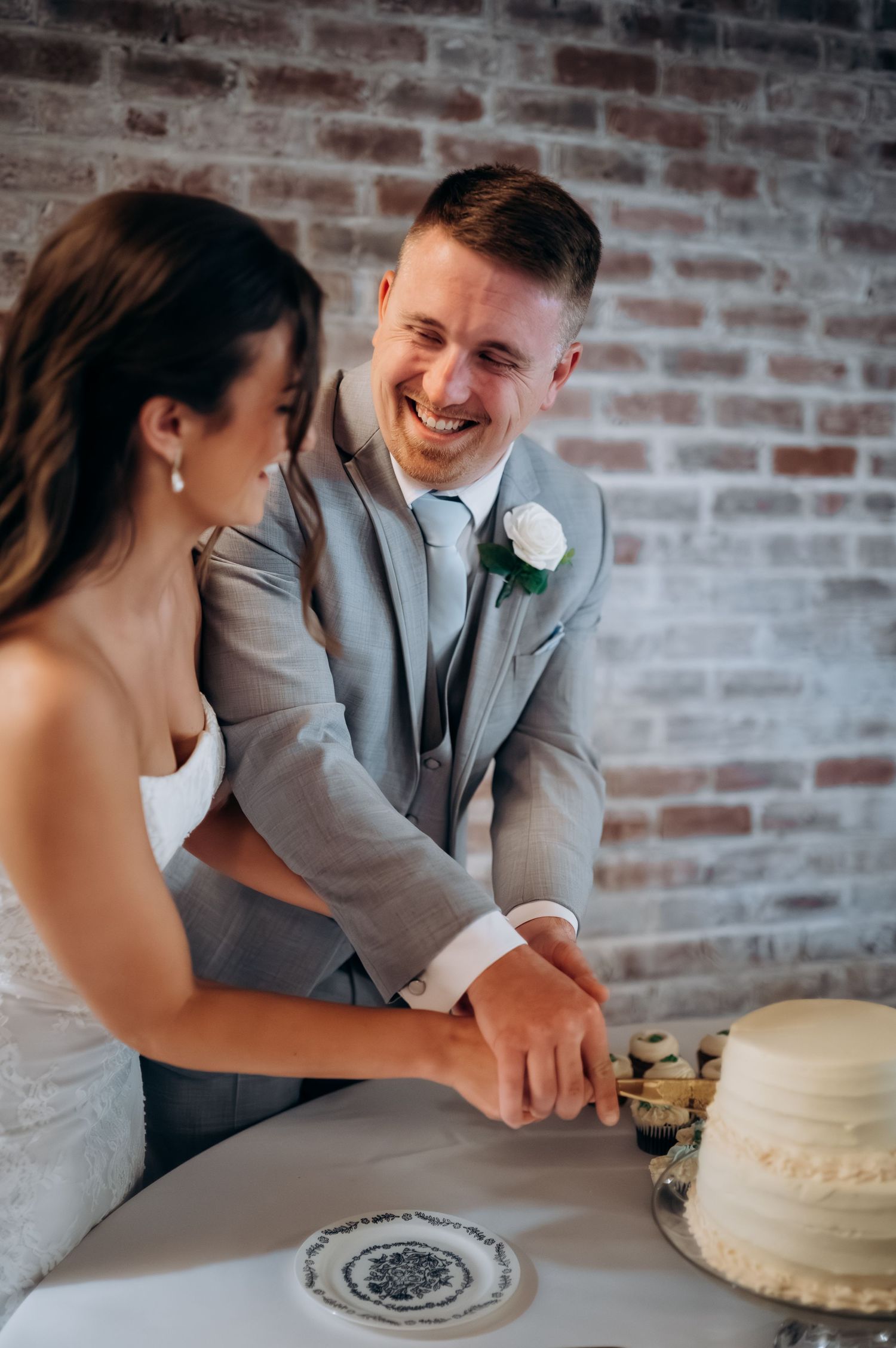 Smiling groom in gray suit cuts white wedding cake with bride against brick wall backdrop.