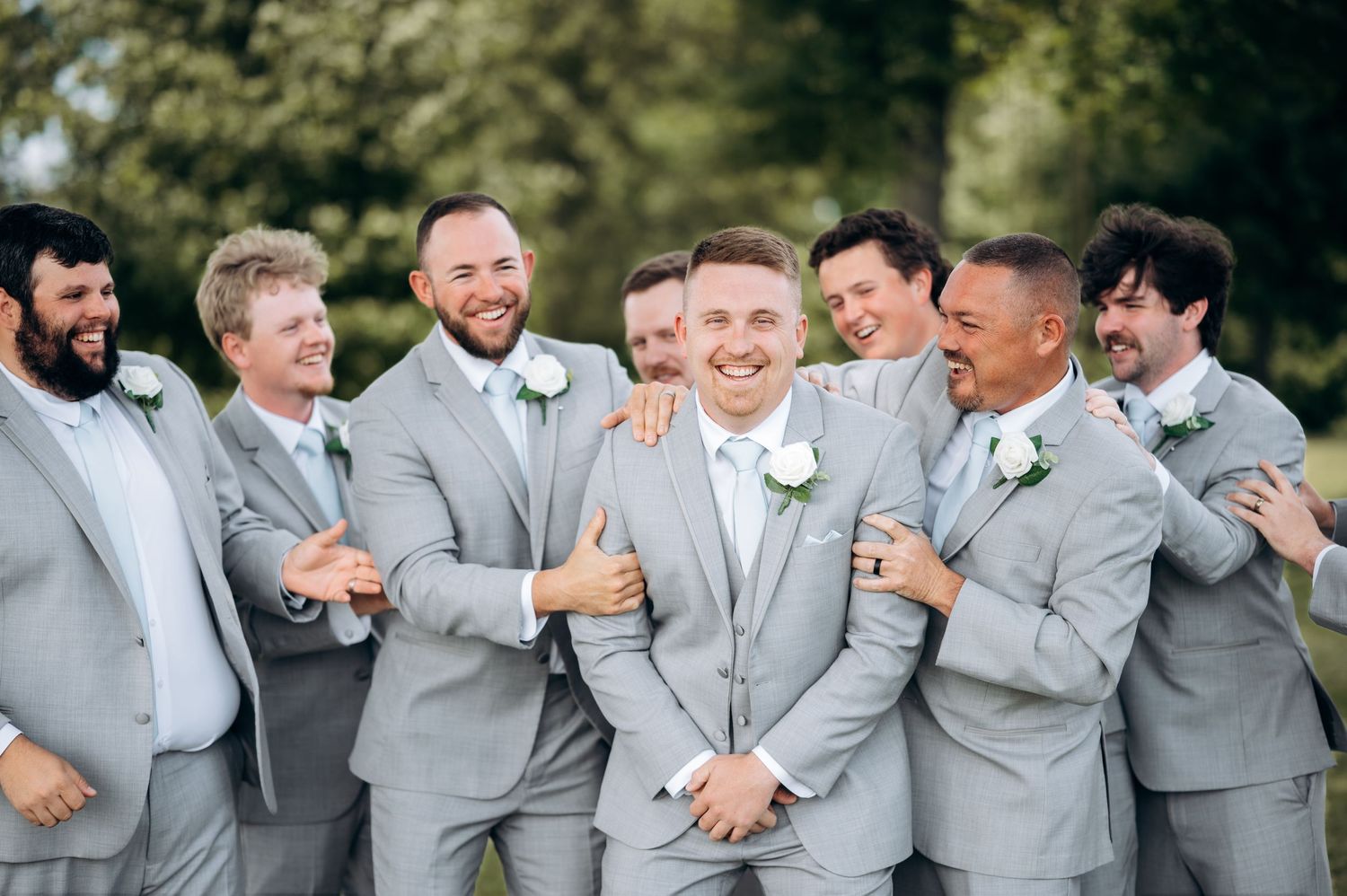 Groom surrounded by smiling groomsmen in matching light gray suits with white boutonnieres at outdoor wedding.