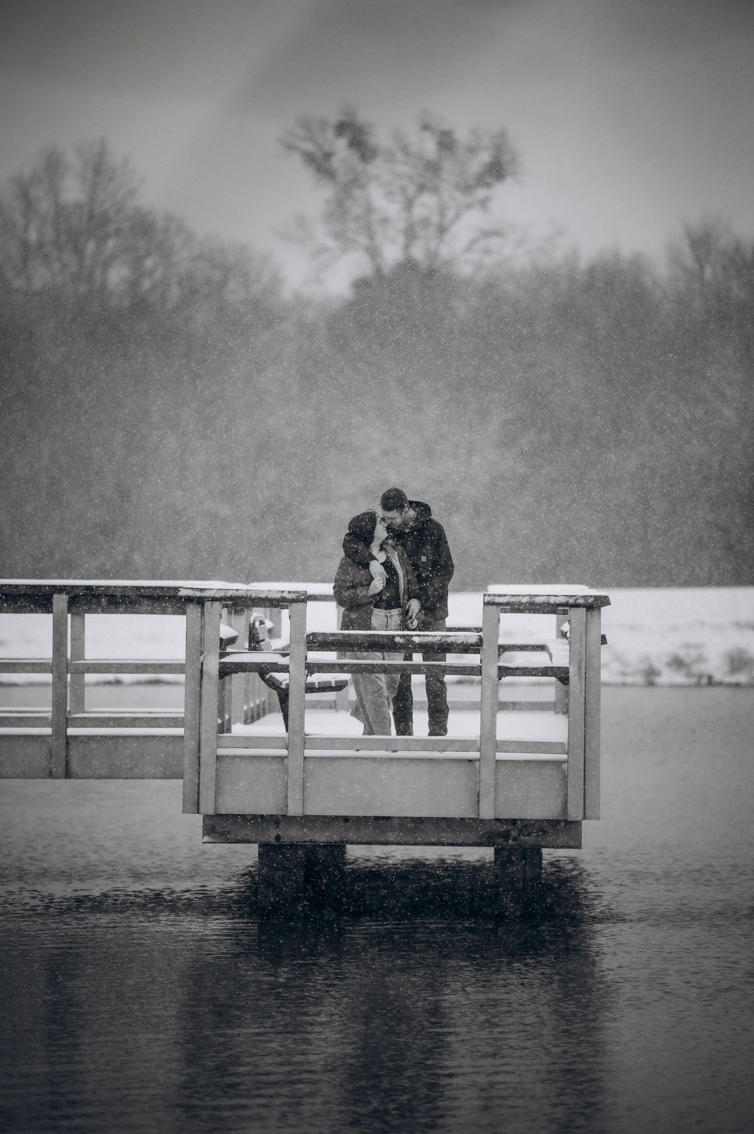 Two figures lean over a wooden dock overlooking a misty lake in black and white photography.