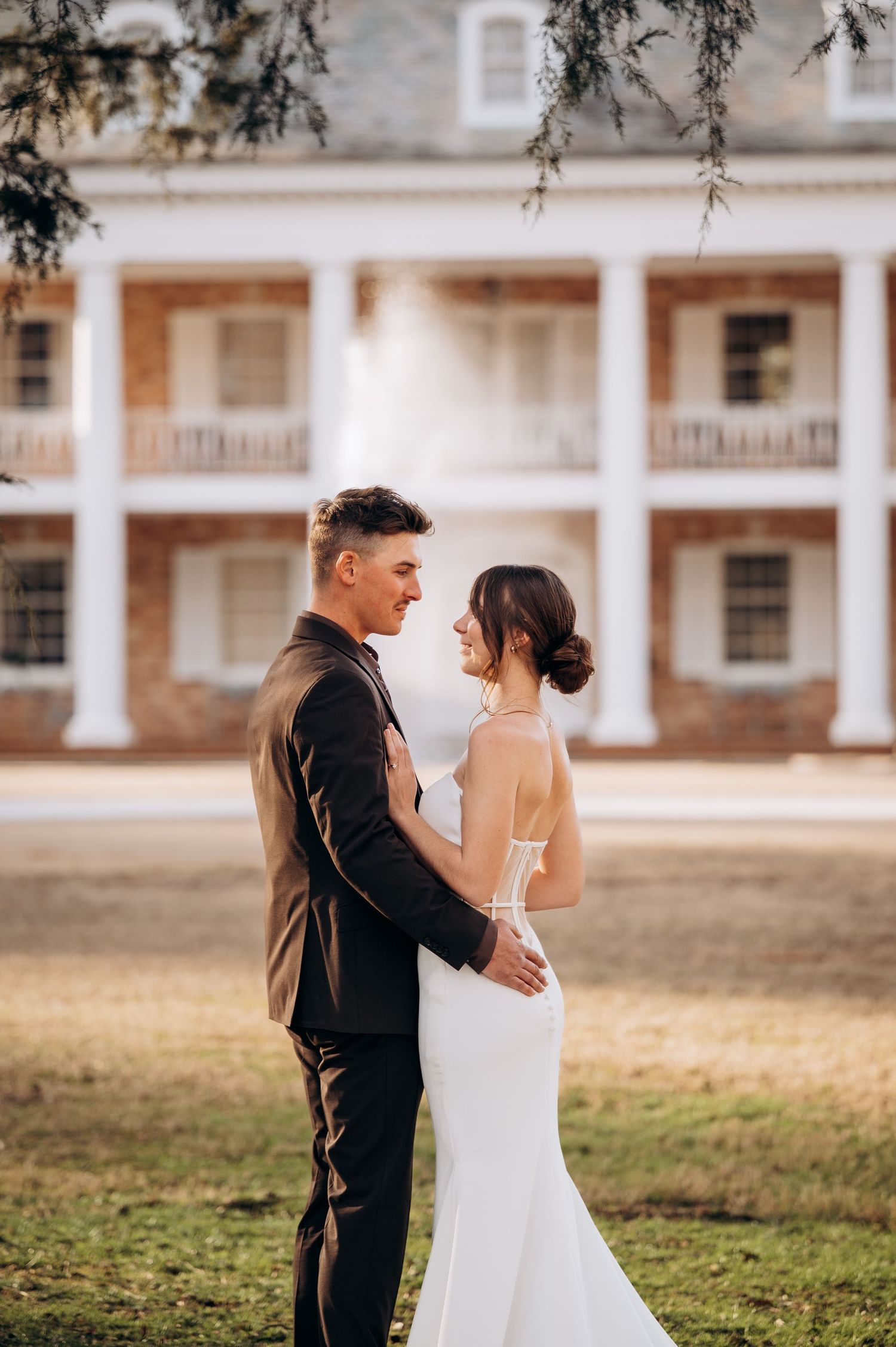 Newlyweds embrace on lawn in front of classic white columned building.