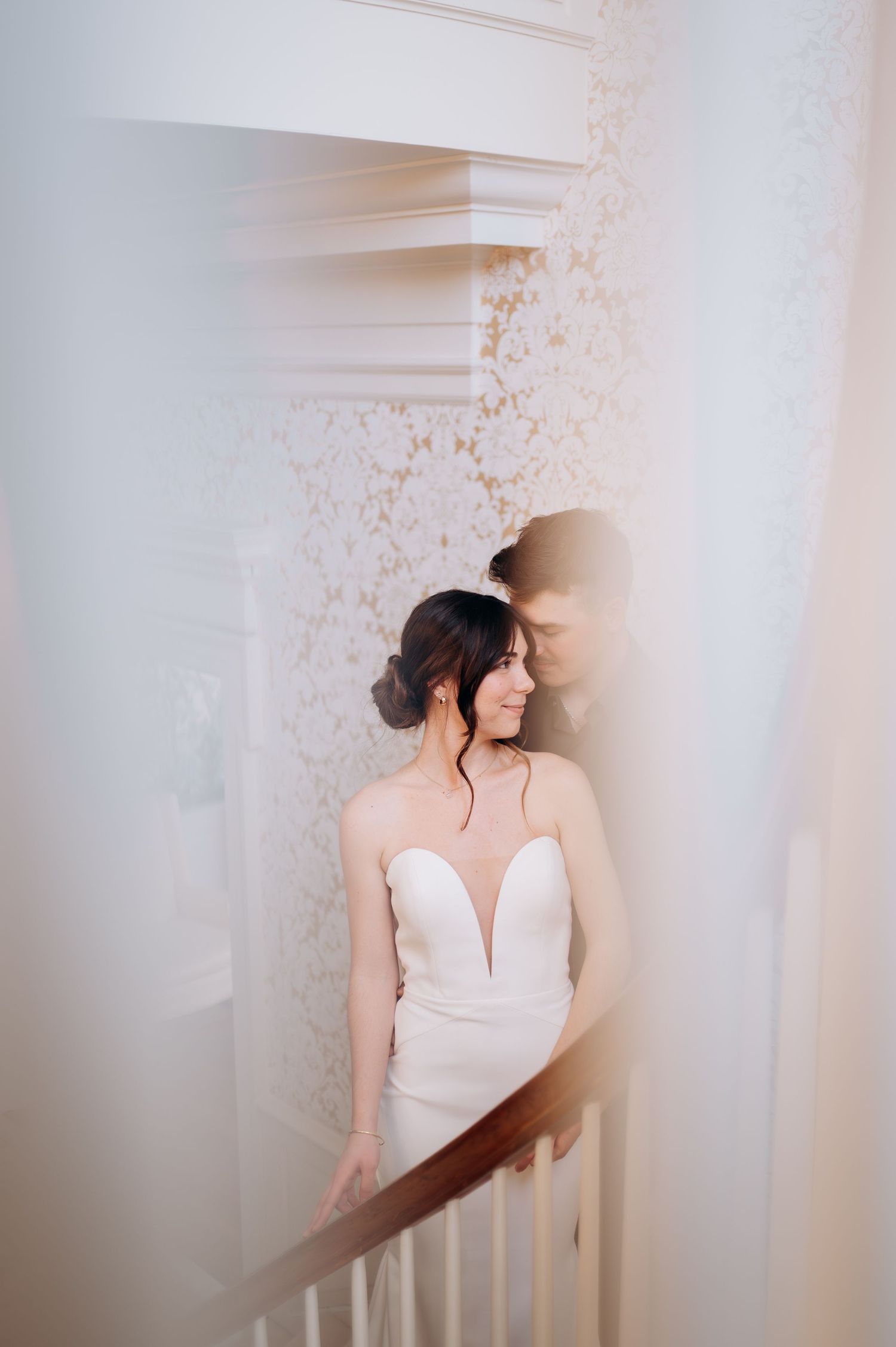 Bride in white gown poses on vintage staircase with delicate floral wallpaper backdrop.