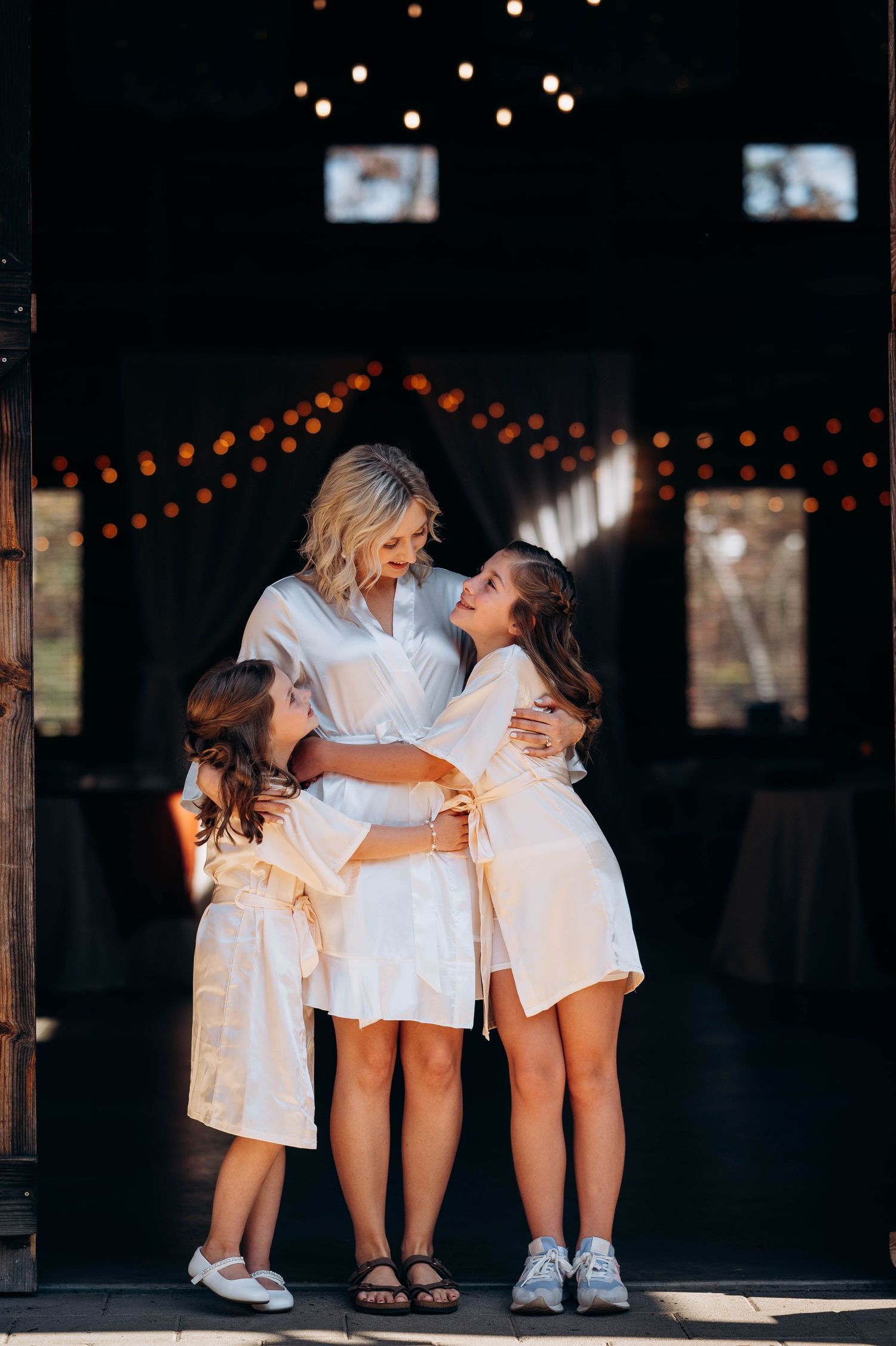 Wedding party in white robes poses playfully under string lights.