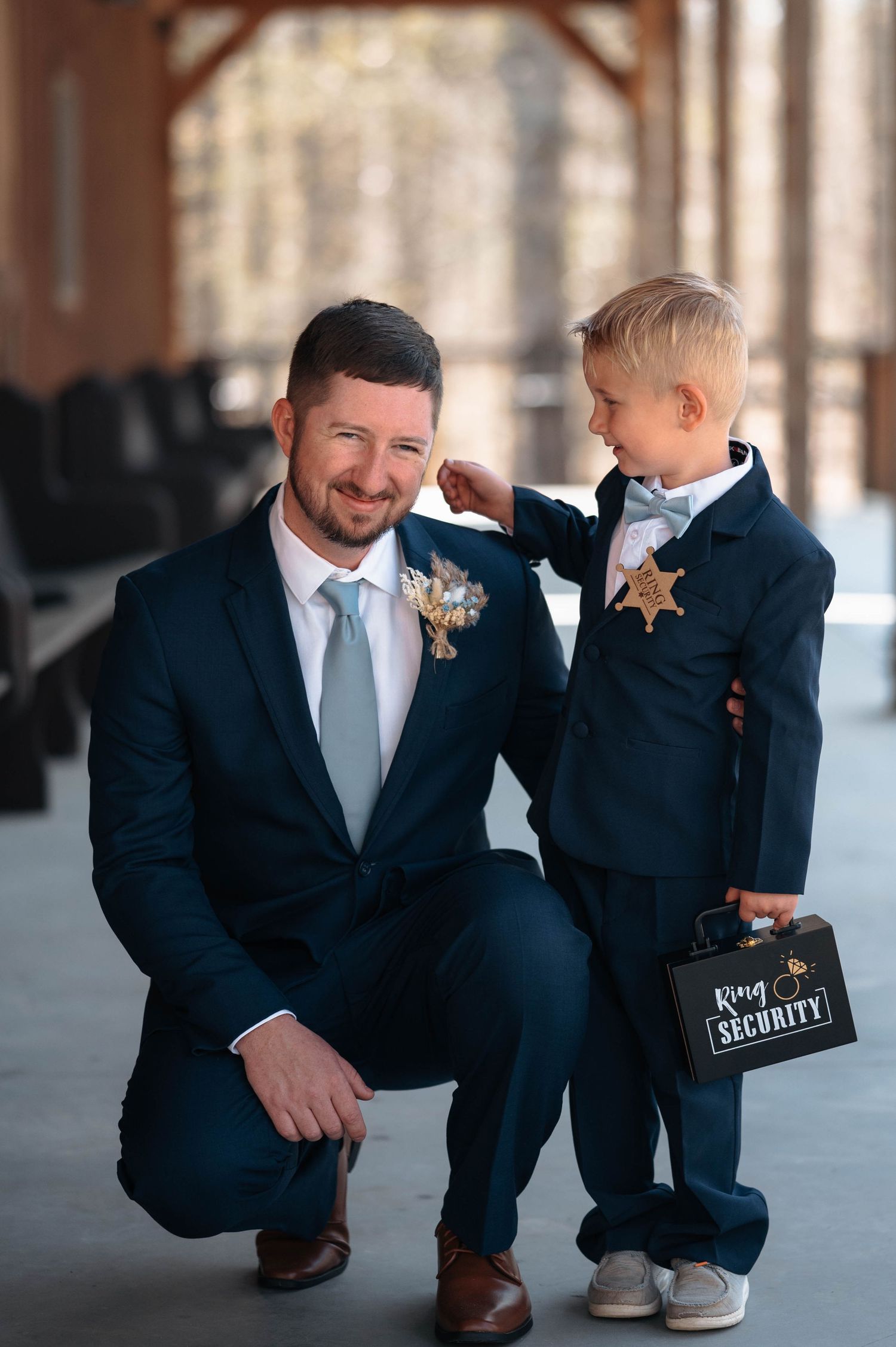 Groomsman in navy suit kneels down to interact with young ring bearer at wedding.