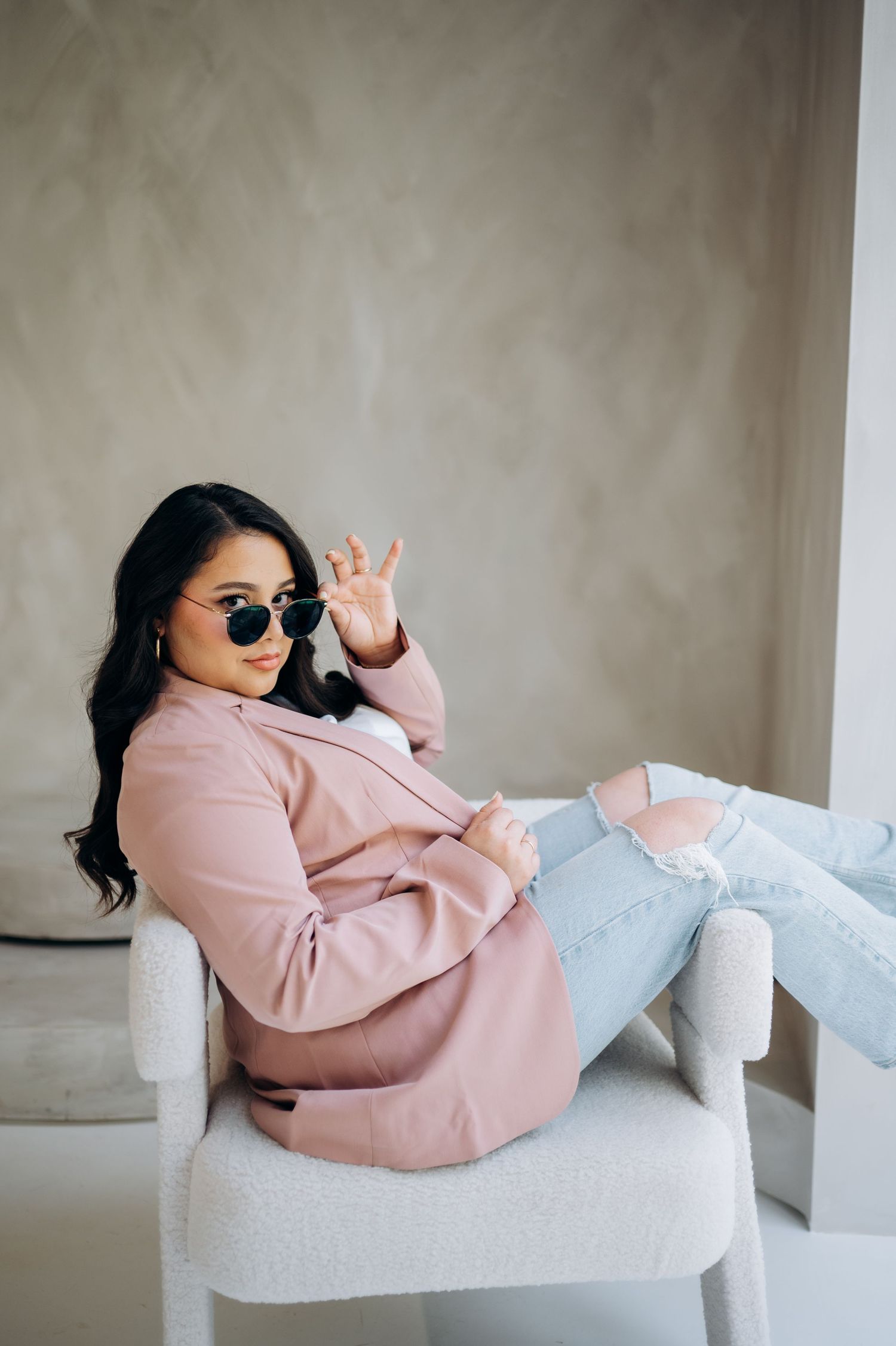 Person in pink jacket and jeans lounges casually on white chair while wearing sunglasses.