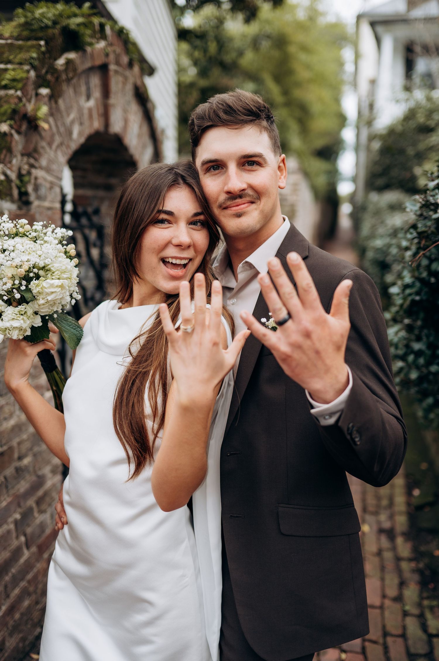 Newly engaged couple showing off their rings with joyful expressions in an outdoor garden setting.