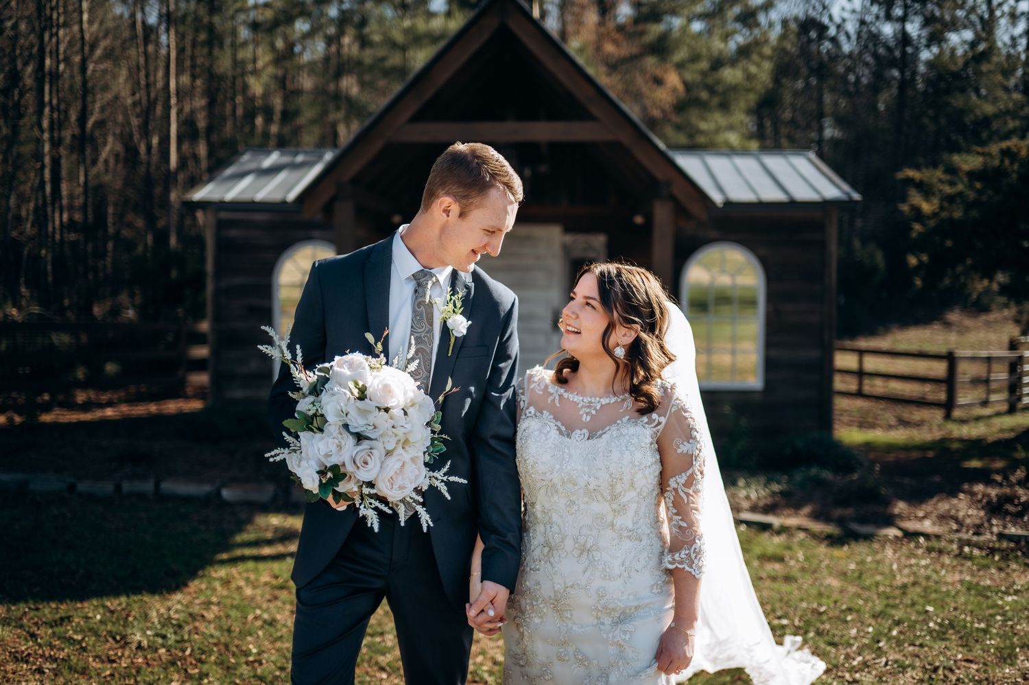 Bride and groom holding white bouquet smile at each other outdoors near rustic wooden chapel on their wedding day.