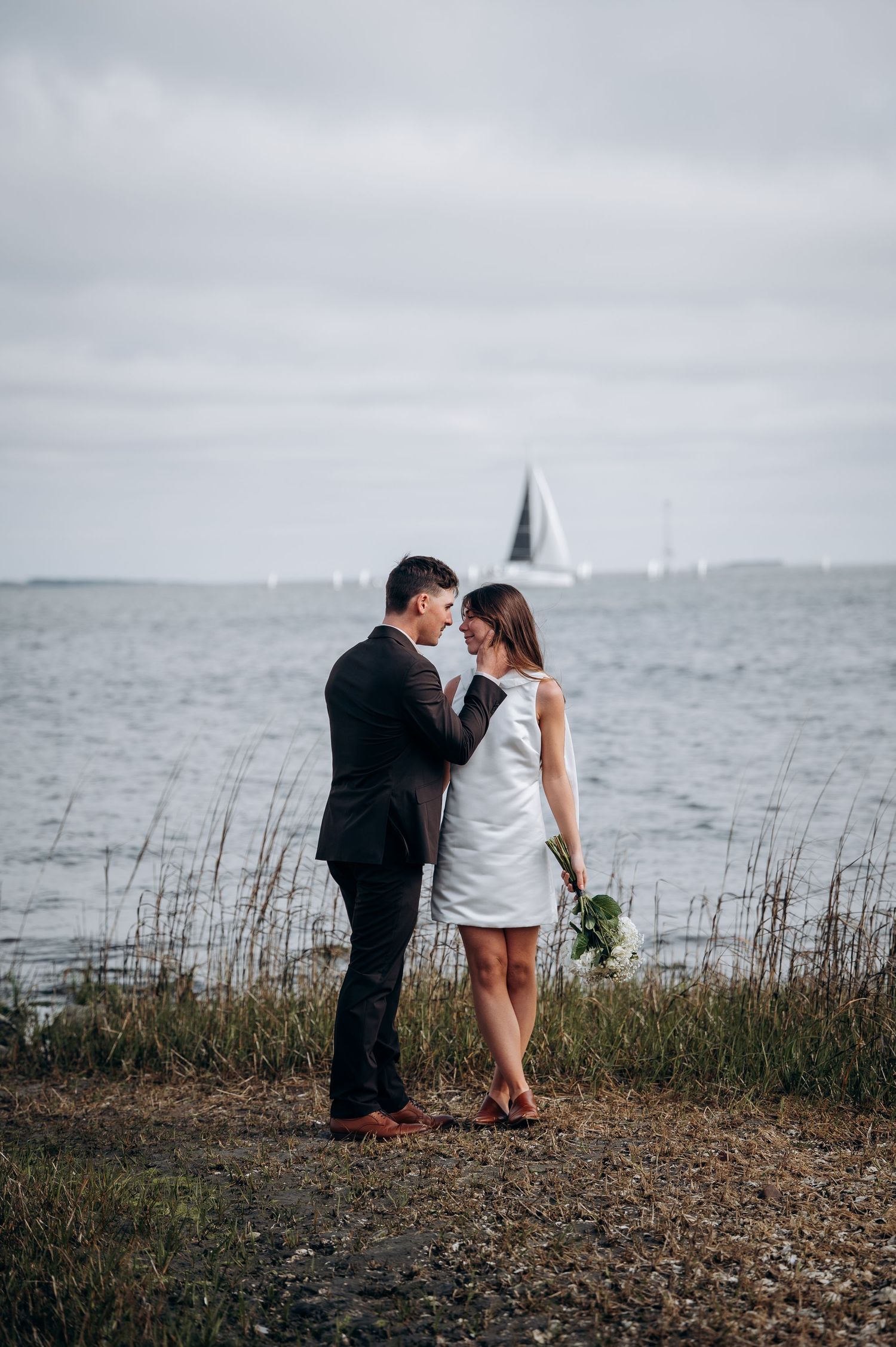 Couple in wedding attire standing by the waterfront, woman holding white flowers, sailboat visible in background.