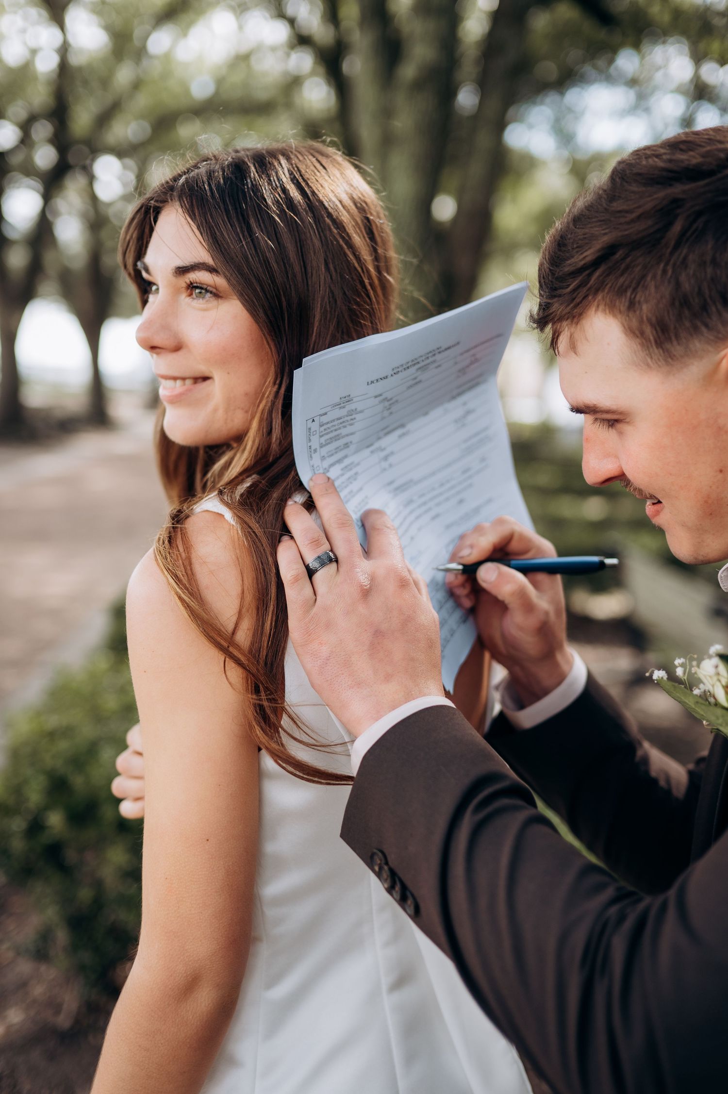 Groom signing marriage license using bride's back as a writing surface outdoors at wedding ceremony.