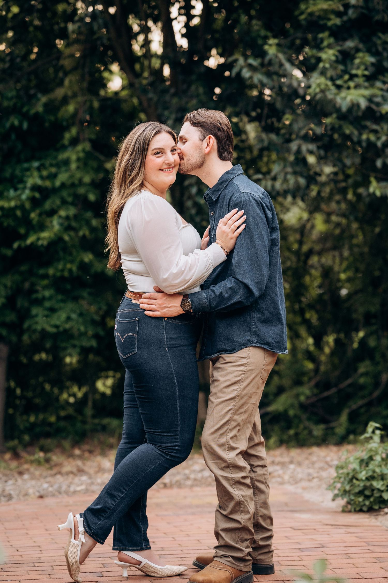 Couple embracing on brick path, man kissing woman on cheek while she smiles, surrounded by lush green trees.