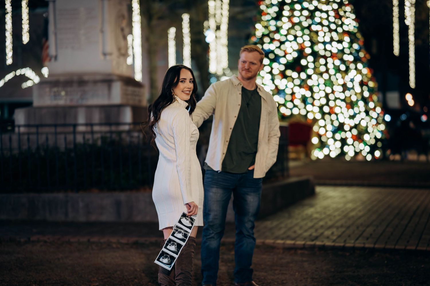 Couple holding ultrasound photos in front of a lit Christmas tree at night for a festive pregnancy announcement.