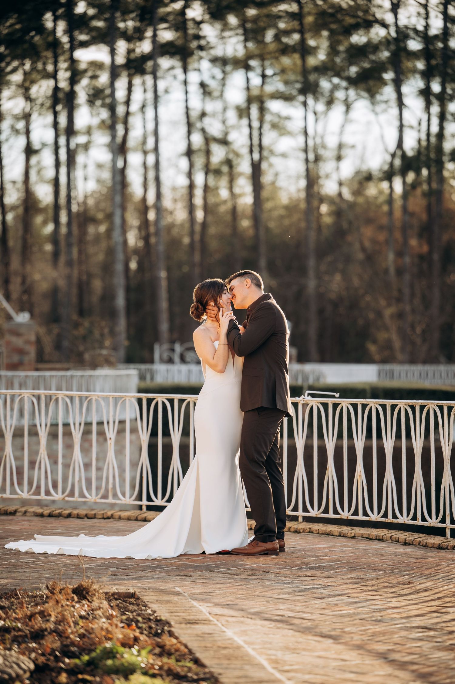 Newlyweds share a tender kiss on a brick terrace with decorative white iron fencing and pine trees.