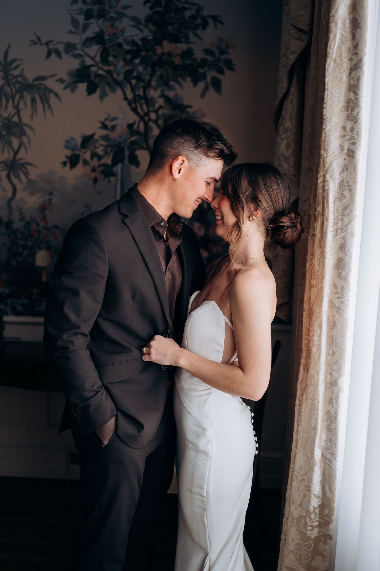 Bride and groom share an intimate moment near a window with floral wallpaper, groom in dark suit, bride in white gown.