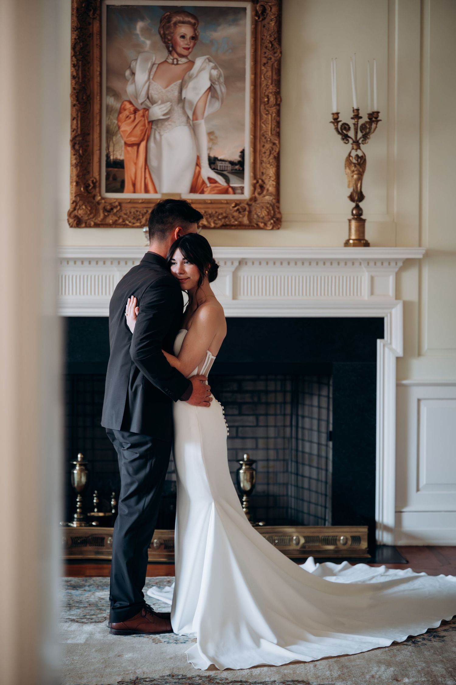 Bride in fitted white gown with train embracing groom by ornate fireplace portrait.