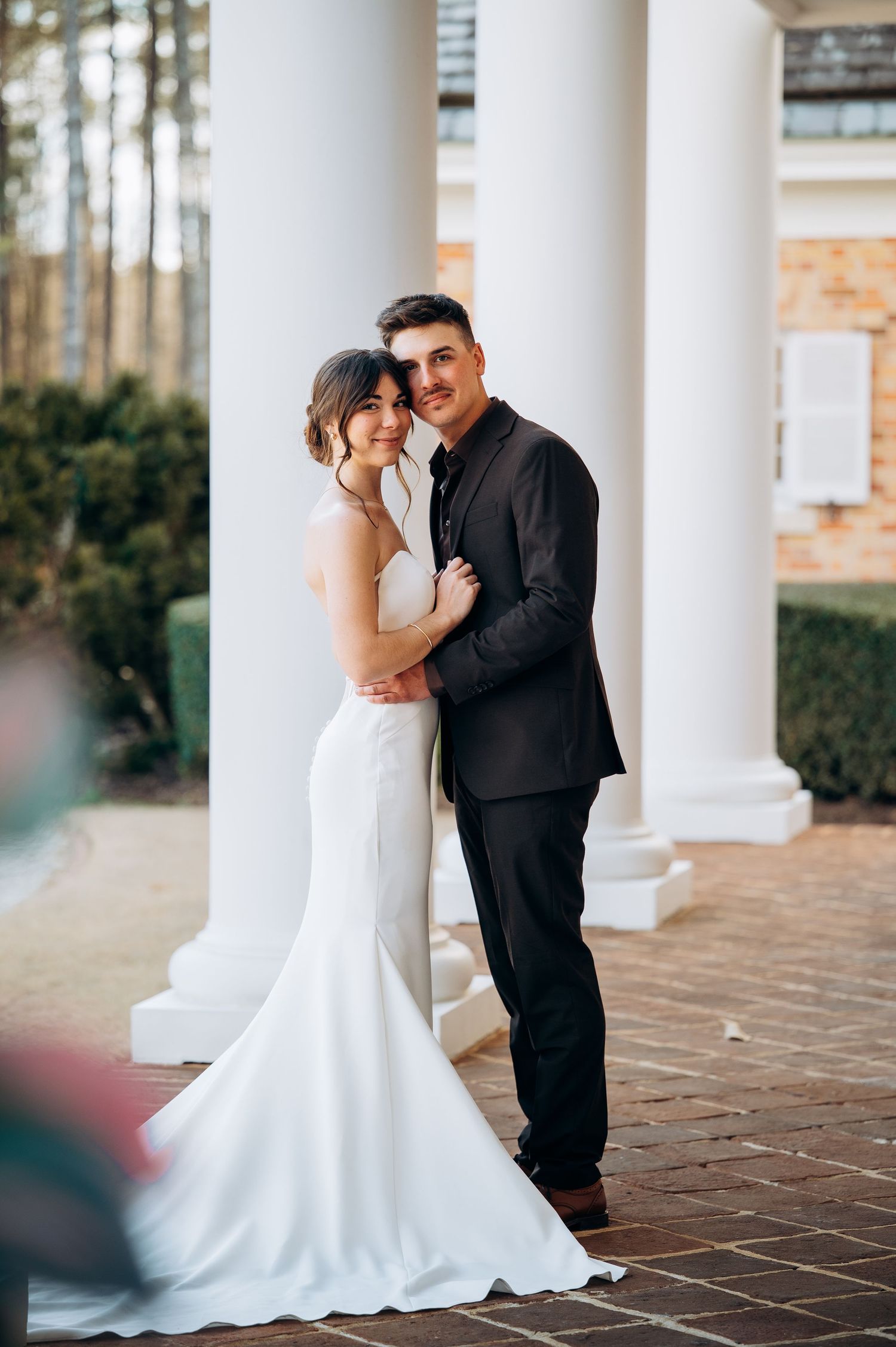 Wedding couple shares intimate moments between elegant white columns on brick patio.