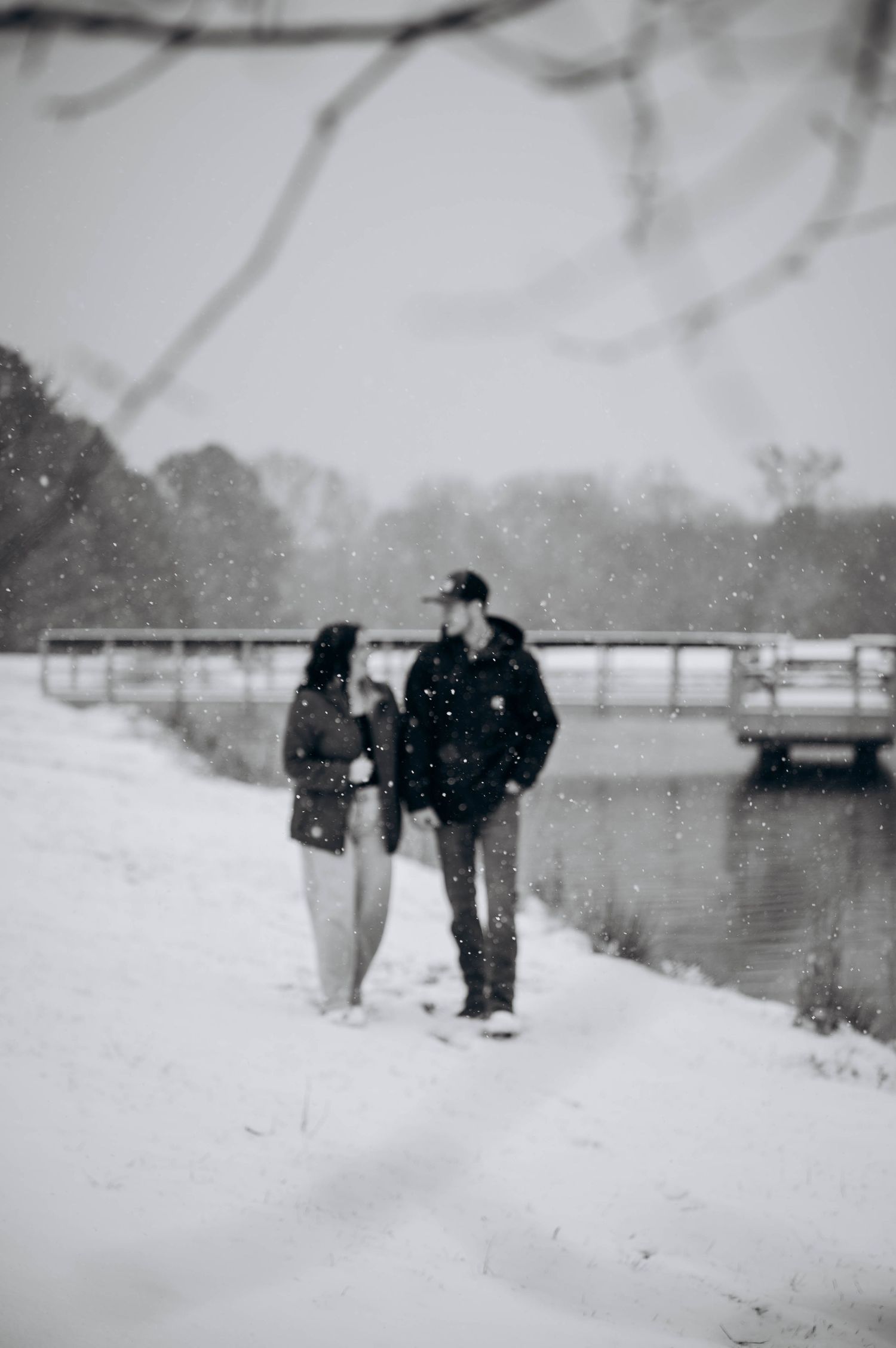 A couple stands together in the snow by a lake with a bridge in the background on a winter day.