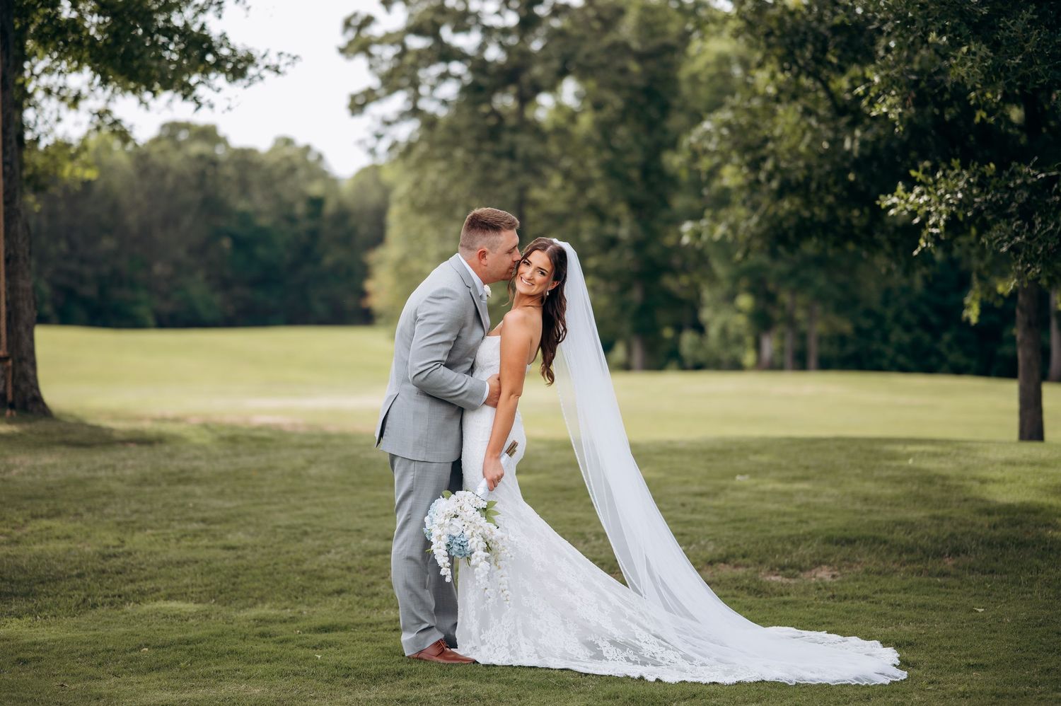 Bride and groom share a romantic kiss on a lush green lawn surrounded by trees on their wedding day.
