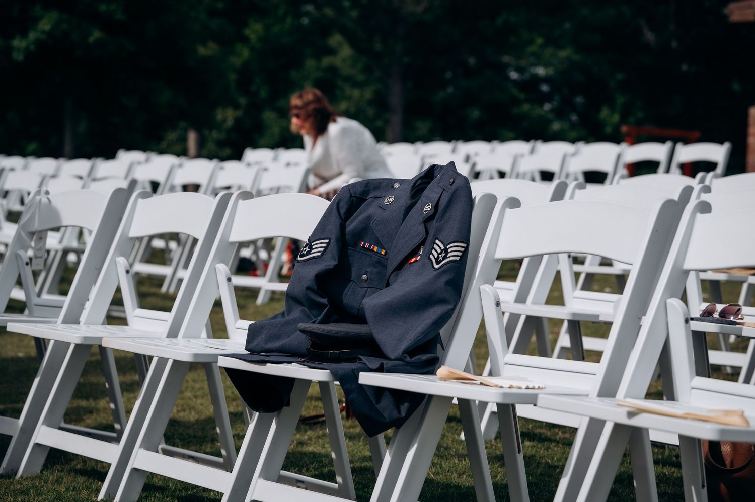 Military jacket draped over white folding chair at outdoor wedding ceremony setup on grass.