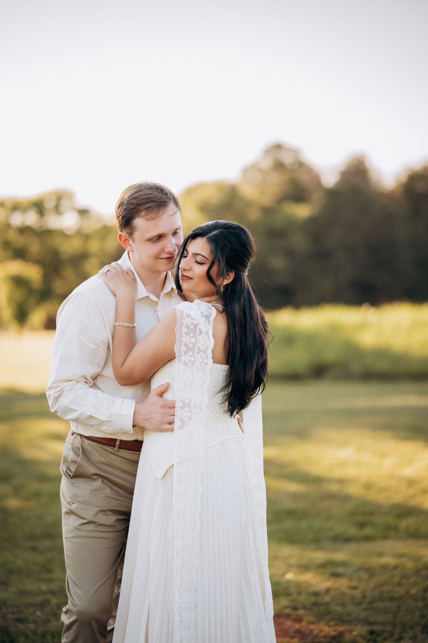 A couple embraces in a sunlit field, the bride in a white lace dress and groom in khaki, sharing a romantic outdoor moment.