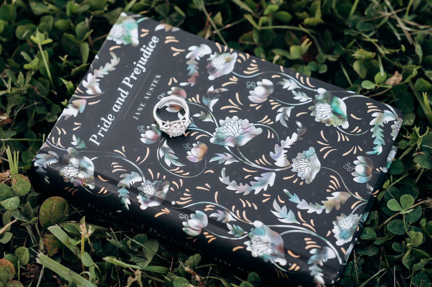 Dark floral edition of Pride and Prejudice by Jane Austen with a ring resting on top, placed on green grass.