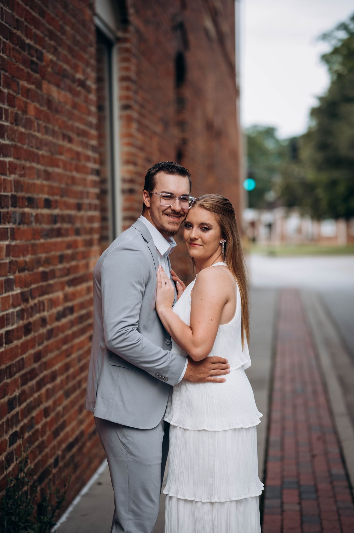 Couple in white and grey formal attire embracing against rustic brick wall backdrop.