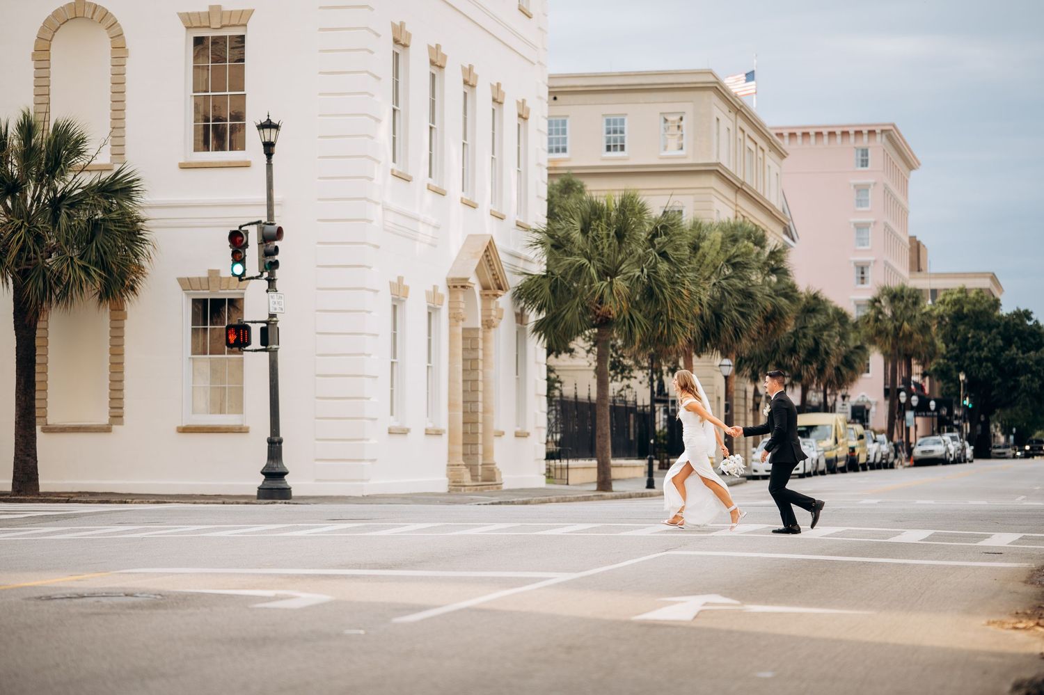 A bride and groom cross a sunny Charleston street lined with palm trees and historic pastel buildings.