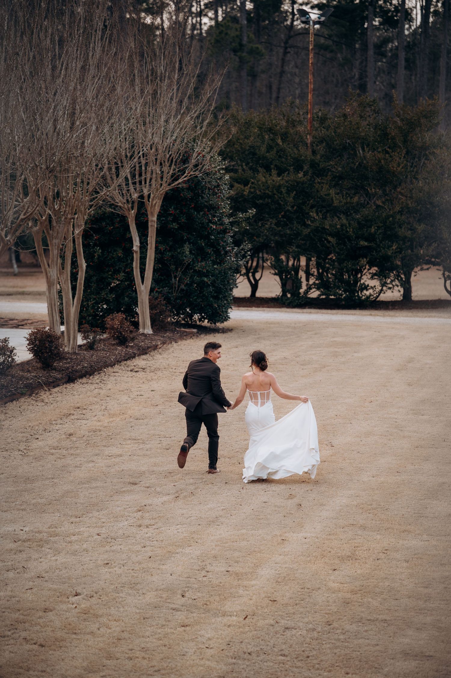 A bride and groom hold hands while walking across a sandy path surrounded by trees during their outdoor wedding.