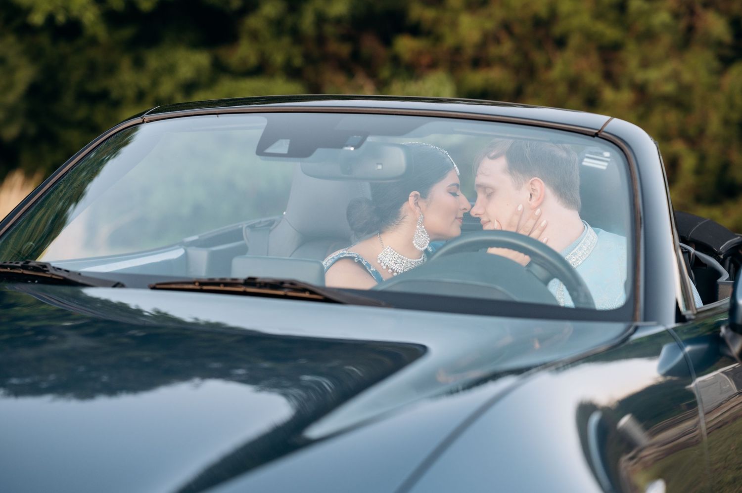 Couple sharing a romantic moment in a black convertible car, leaning in for a kiss on a sunny day.