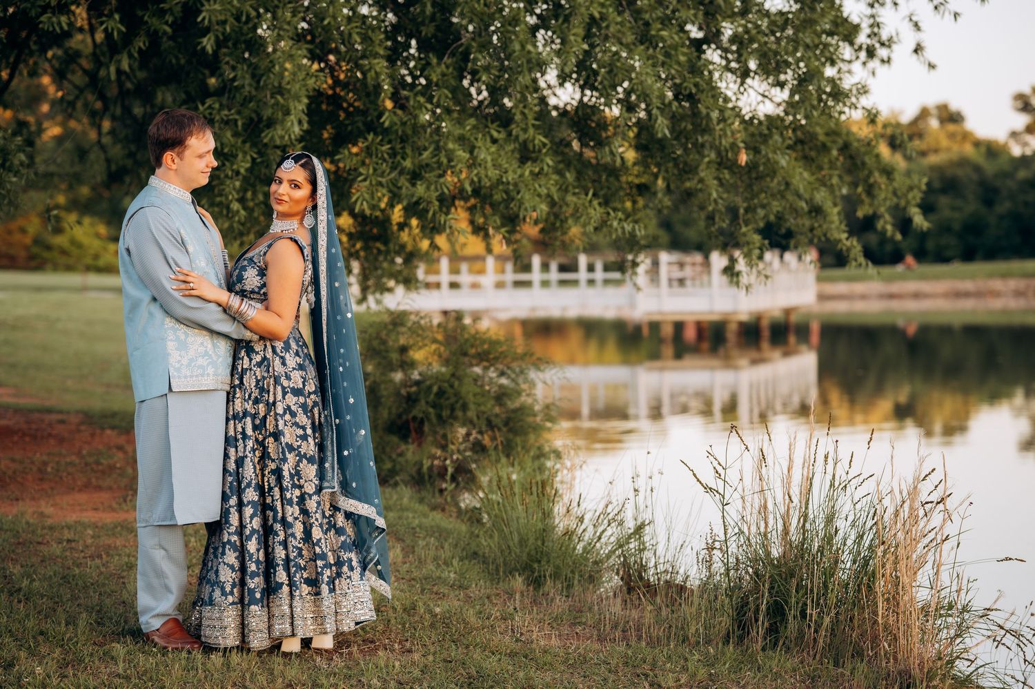 Couple in Indian wedding attire poses by a serene pond surrounded by lush greenery at golden hour.