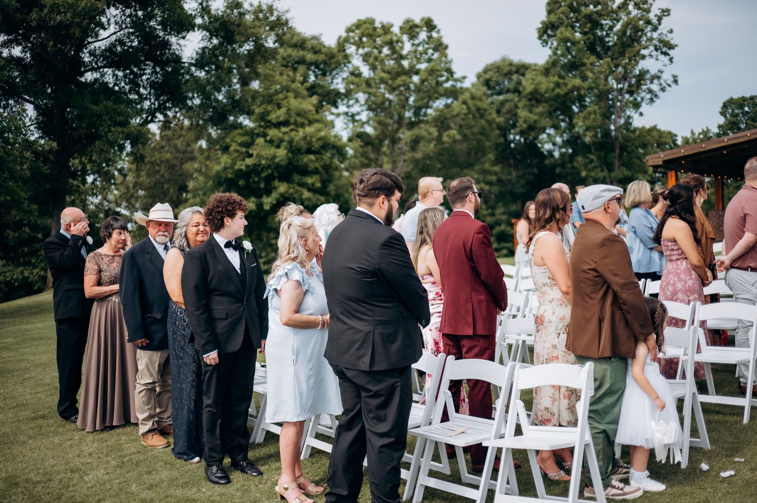 Wedding guests standing and mingling at elegant outdoor ceremony with white chairs on manicured lawn.